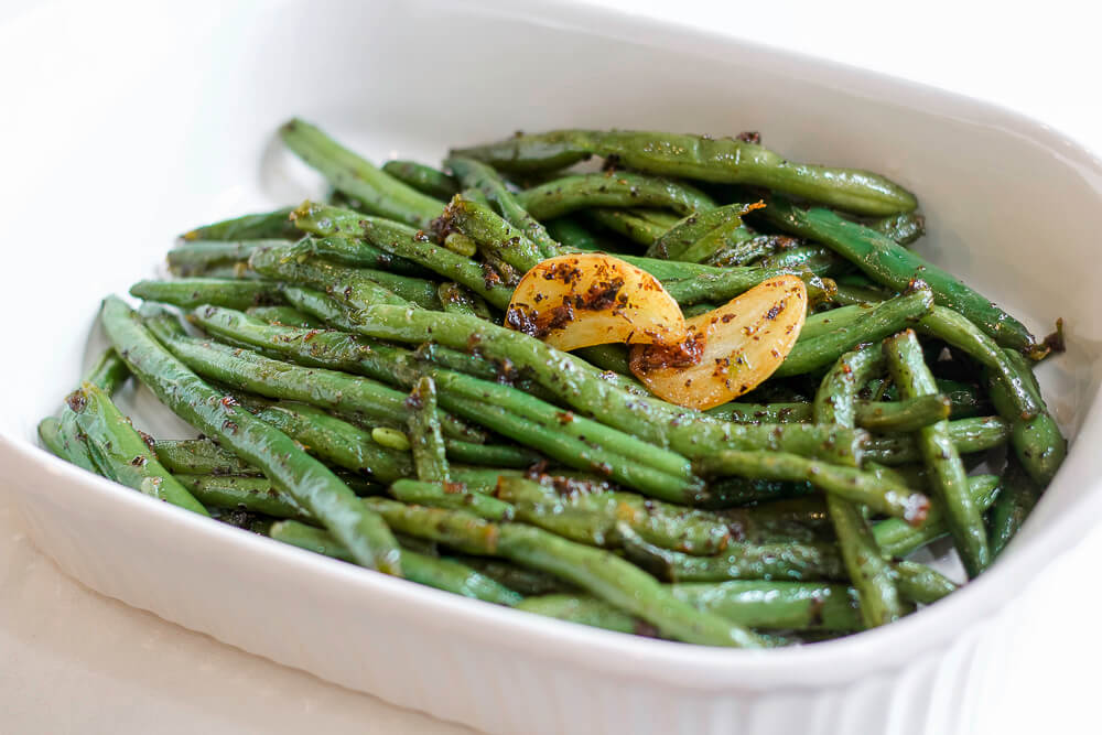 A white serving dish filled with sautéed fresh green beans, garnished with roasted garlic slices.