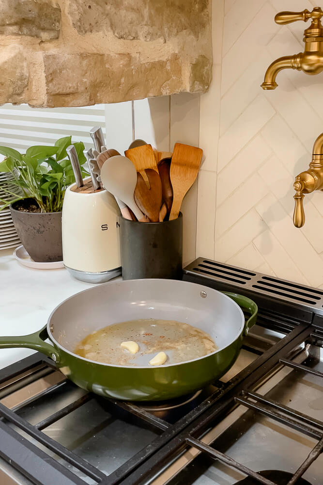 A green pan on the stove with garlic cloves sautéing in oil, preparing for fresh green bean