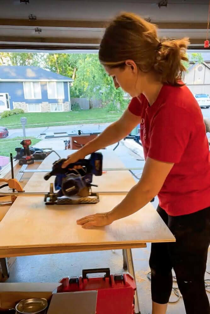 Cutting a piece of plywood with a circular saw, guided by a clamped straight edge for precision, to create custom pull-out shelving.