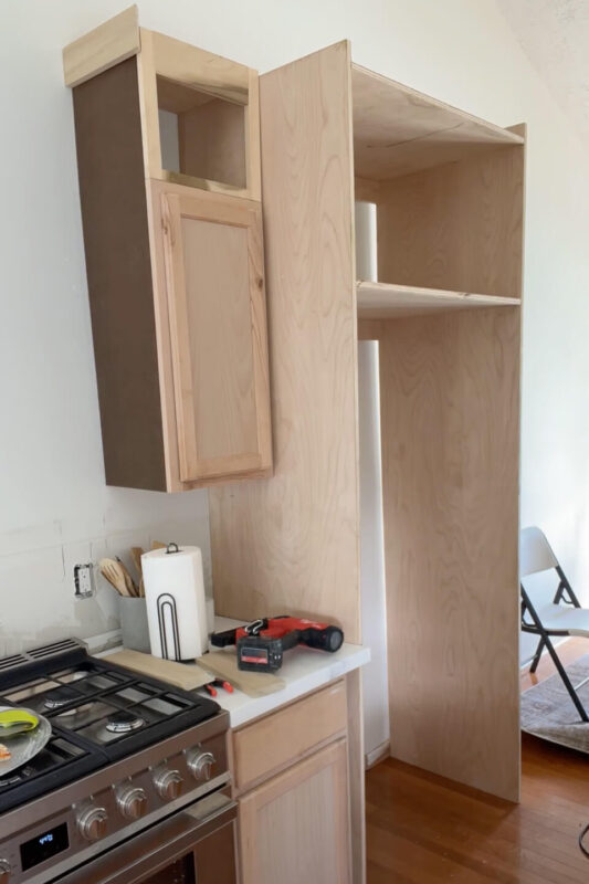 Unfinished DIY refrigerator cabinet next to other kitchen cabinets.