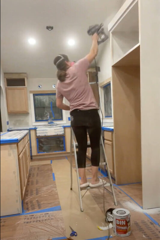 Woman using paint sprayer to prime DIY kitchen cabinets.