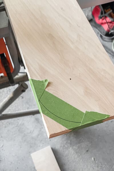 Green painter’s tape marking a curved cut on the corner of a wood board for DIY Murphy bed