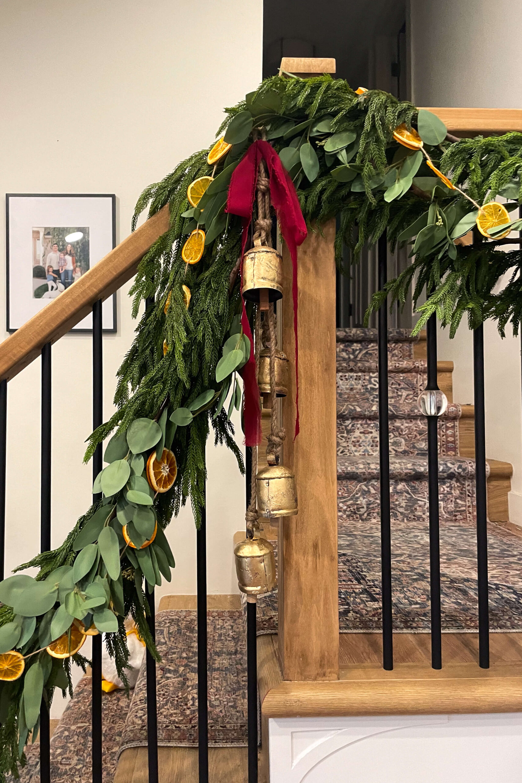 Red bows, gold bells, orange slice garland, and realistic, faux pine garland on a Christmas staircase.