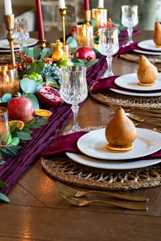 Burgundy Thanksgiving tablescape place setting with a golden pear on a white plate, burgundy napkin, gold flatware, and glass goblets