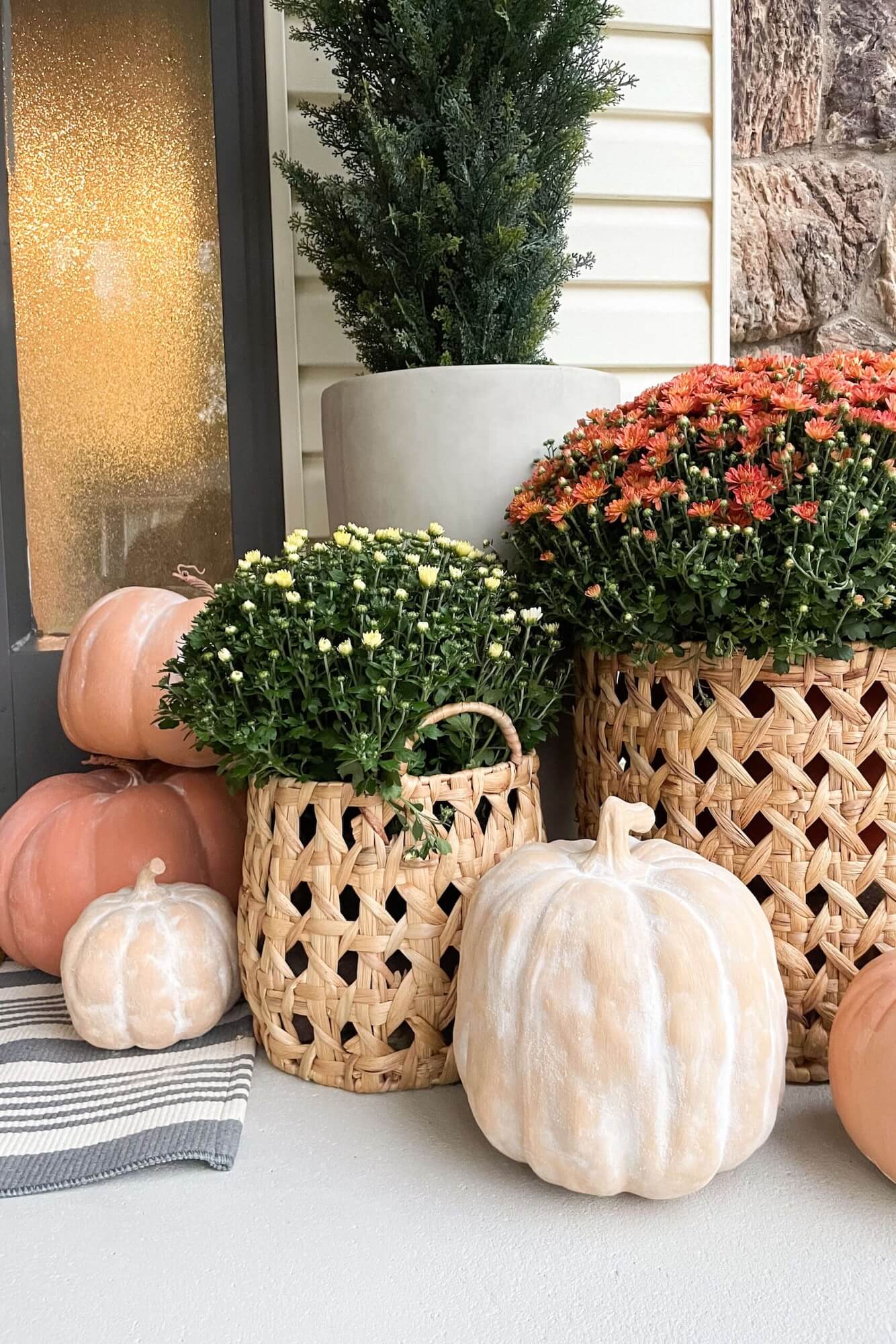 Neutral fall porch styling with woven baskets of mums, stacked pumpkins, and a tall evergreen by the front door.