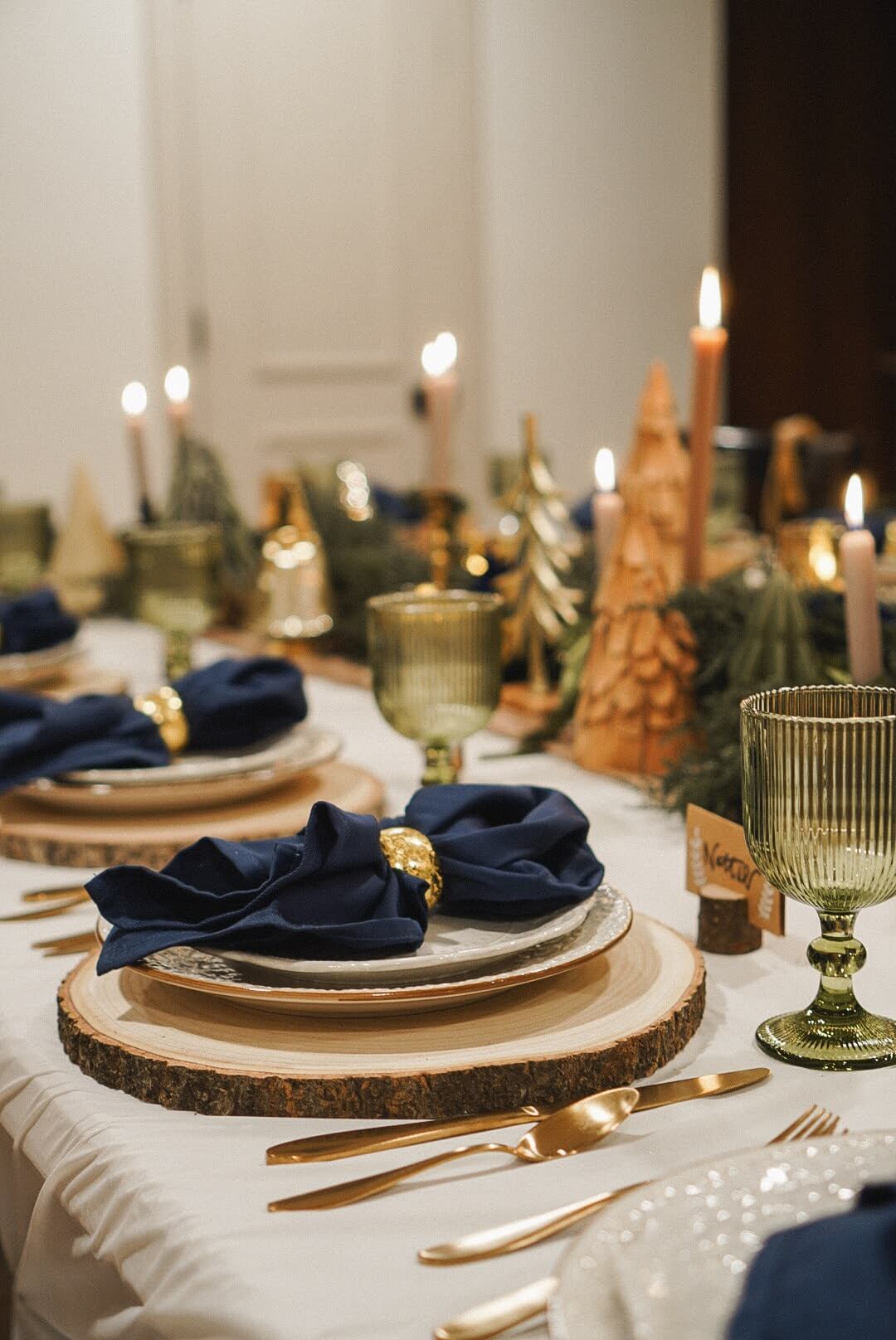 Elegant Christmas tablescape with green goblets, wood slice chargers, and navy blue napkins, surrounded by warm candlelight and holiday decor.