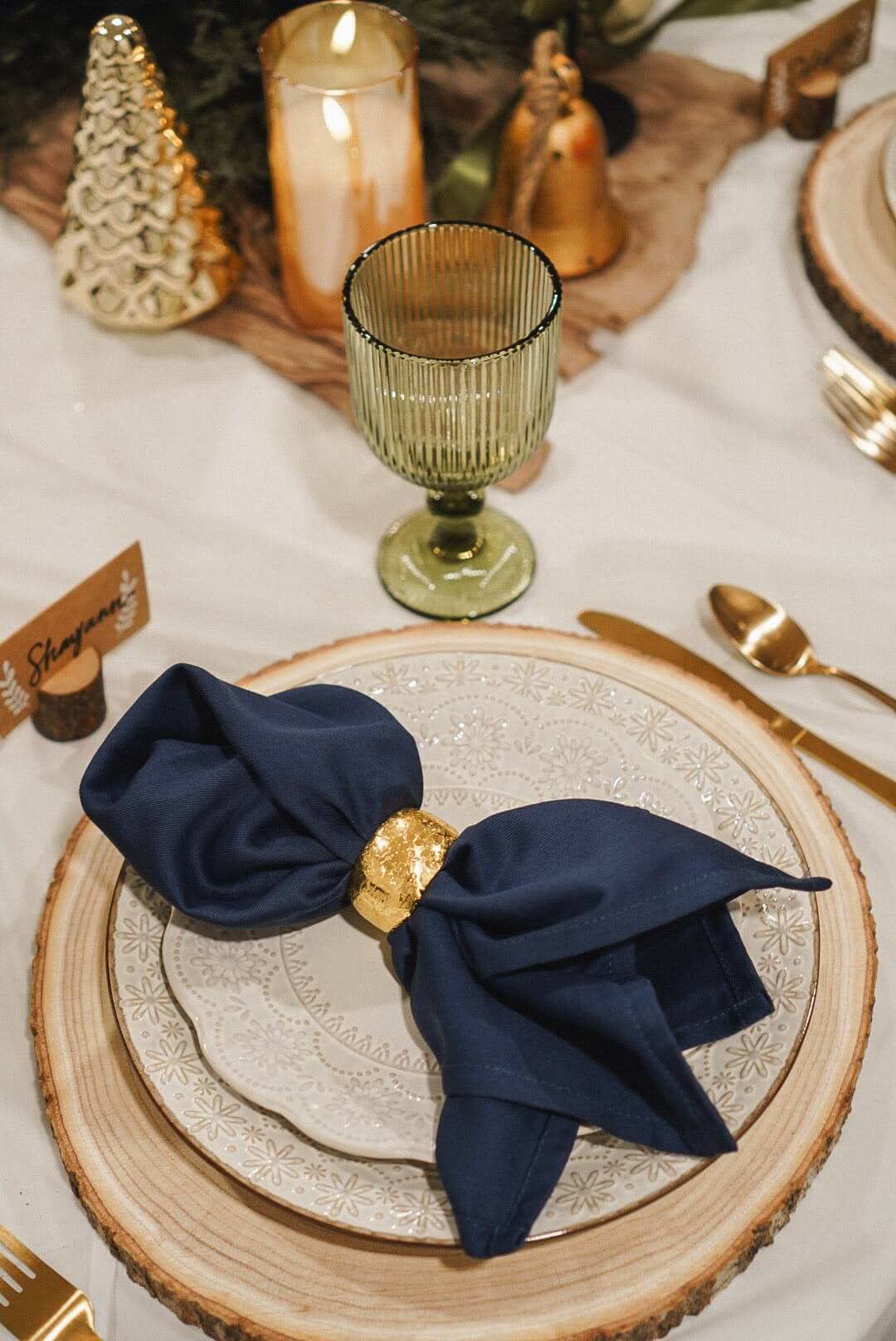 Place setting with a navy blue napkin and gold napkin ring, wood slice charger, and green goblet on a detailed Christmas tablescape.