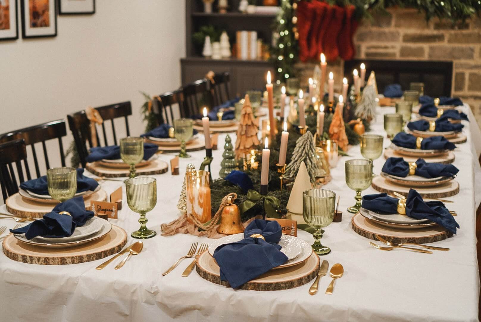 Complete view of a Christmas tablescape with navy napkins, wood slice chargers, and green goblets, surrounded by Christmas trees and candles.