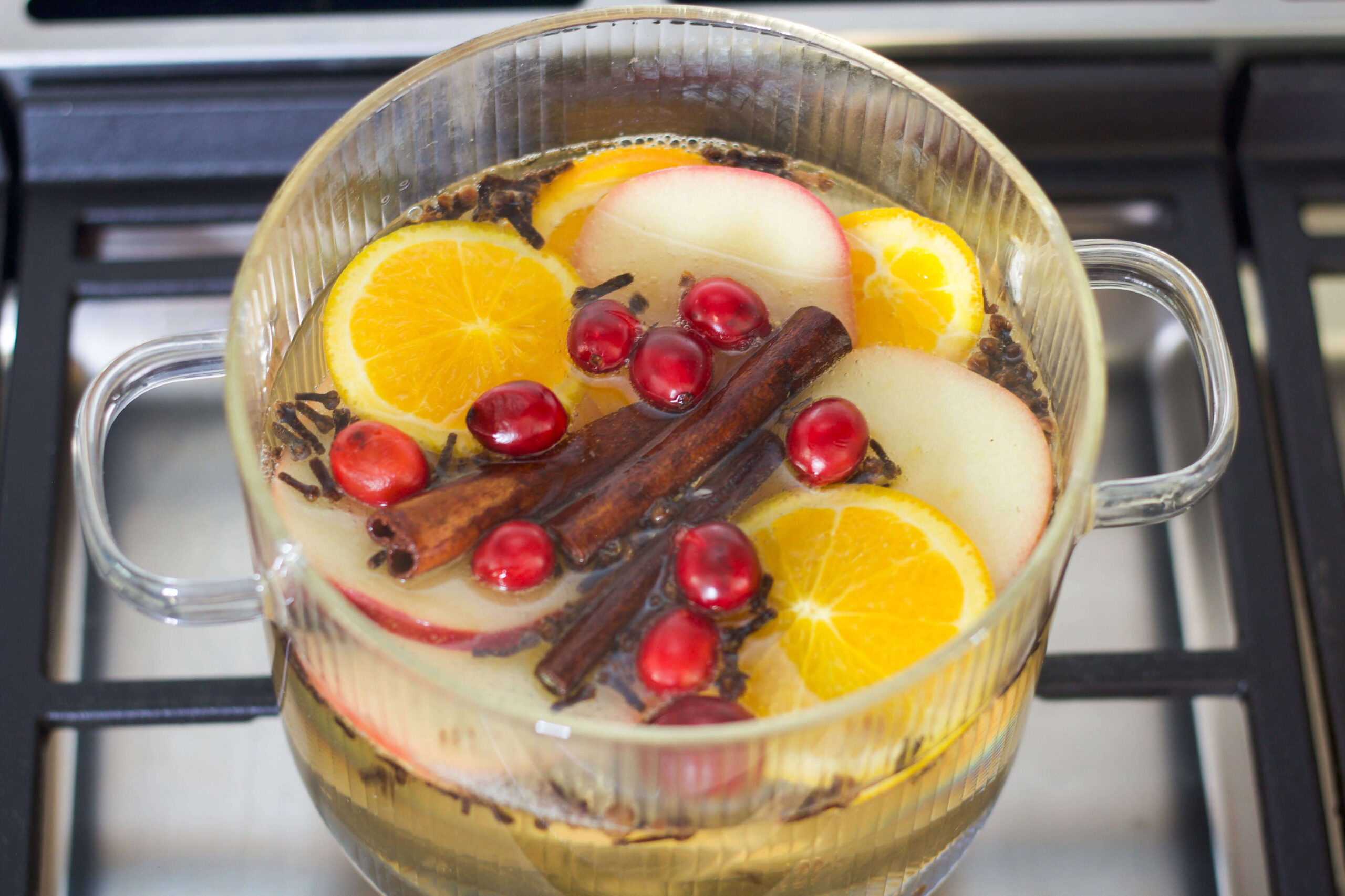 A Christmas simmer pot with orange slices, apple slices, cranberries, and cinnamon sticks simmering in a clear pot on the stove.