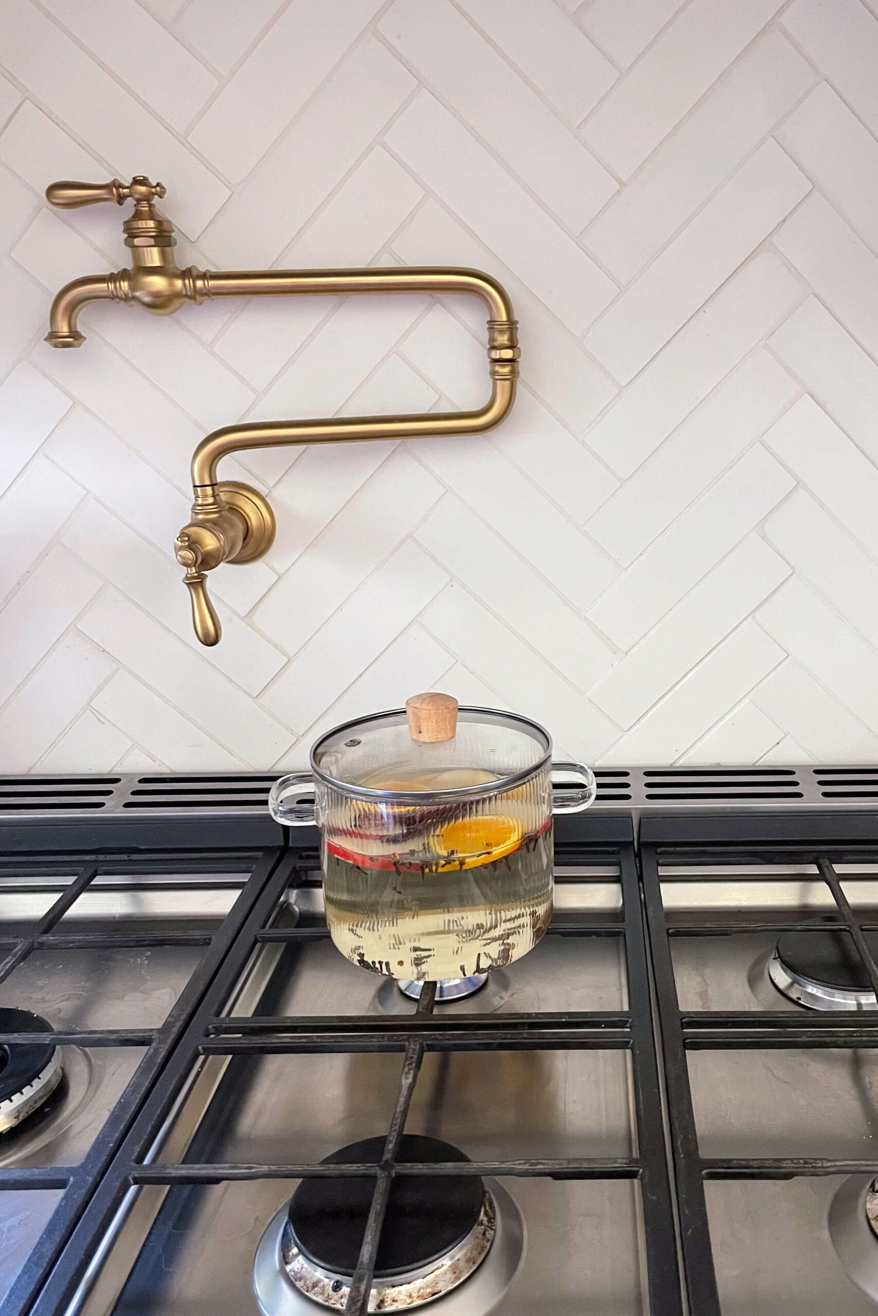 Glass pot with water, apple slices, orange slices, and cinnamon sticks simmering on a stove beneath a brass pot filler with a herringbone tile backsplash.