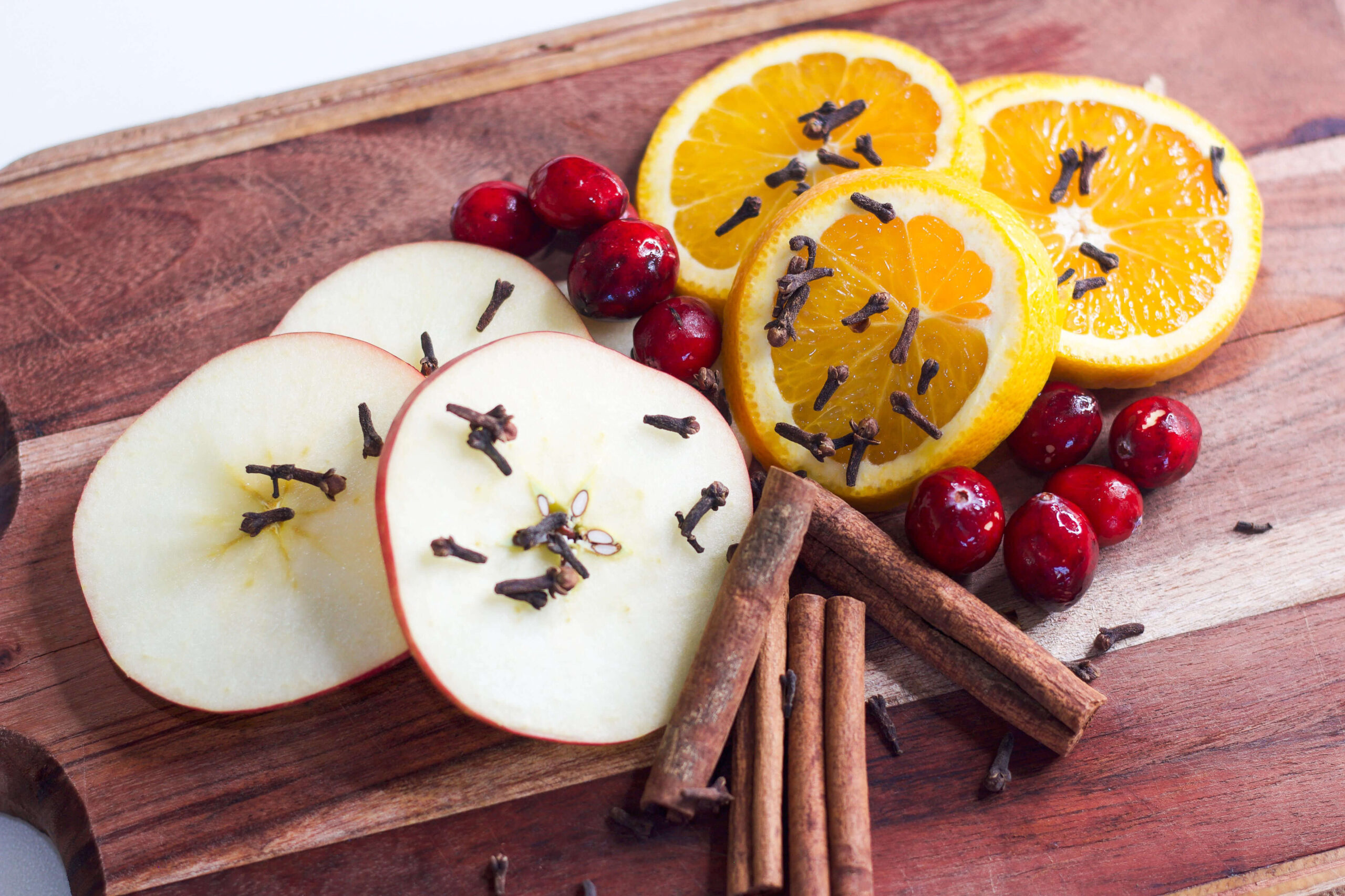 Sliced apples, oranges, and cranberries with cloves and cinnamon sticks arranged on a wooden board for a Christmas simmer pot.