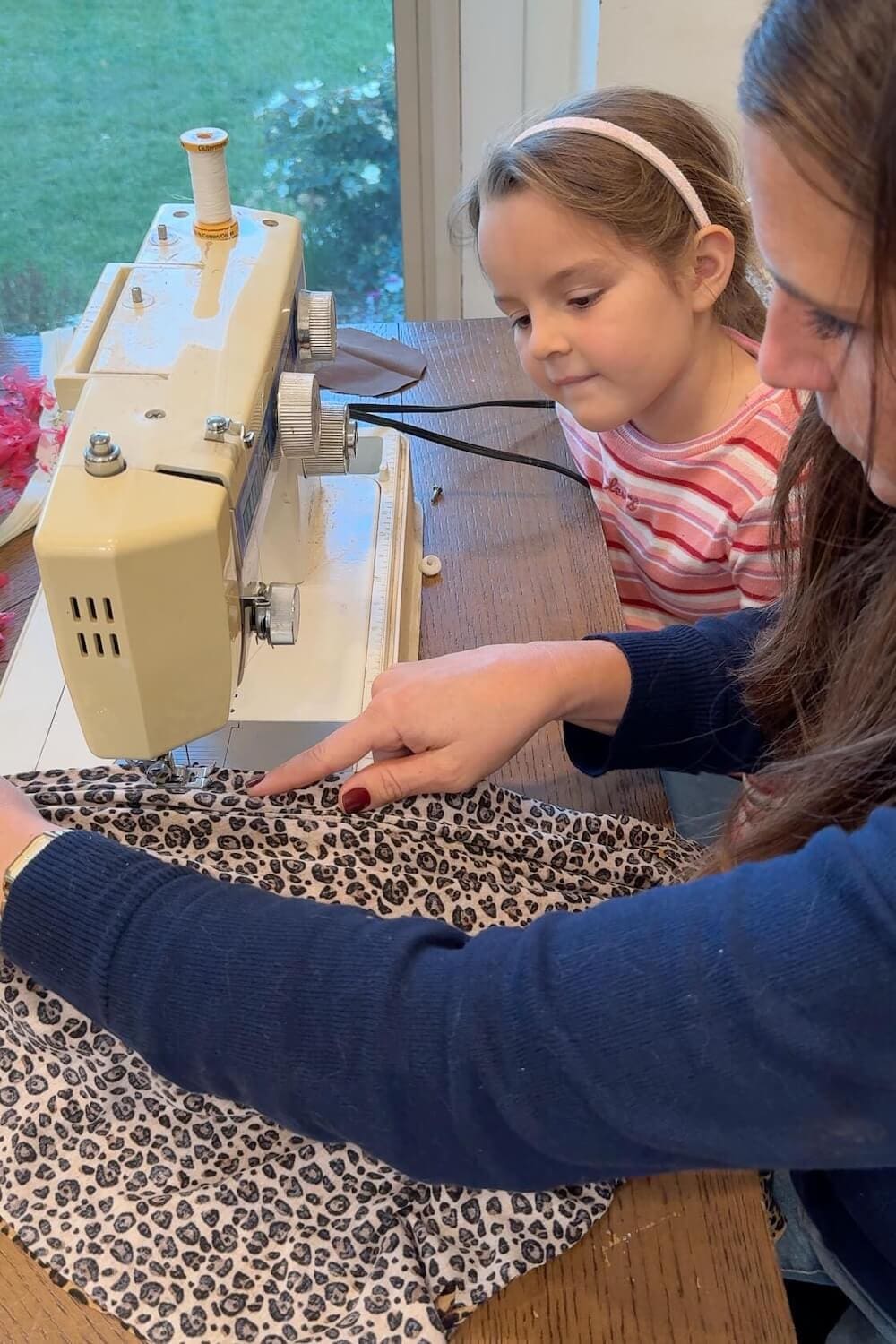Parent guiding a child while hemming a leopard-print tunic on a sewing machine.