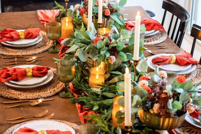 Thanksgiving table decorations from the end of the table: woven placemats, terracotta napkins in gold rings, and a greenery centerpiece with candles