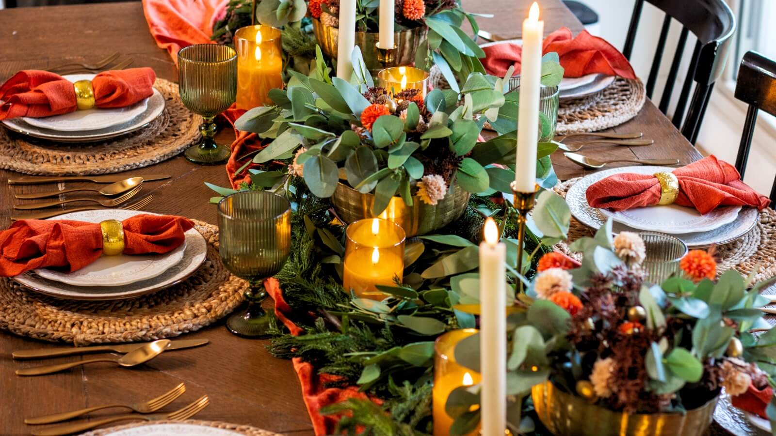 Thanksgiving table decorations with brass bowls of faux mums and eucalyptus, flameless candles, green goblets, and terracotta napkins in gold rings on woven placemats
