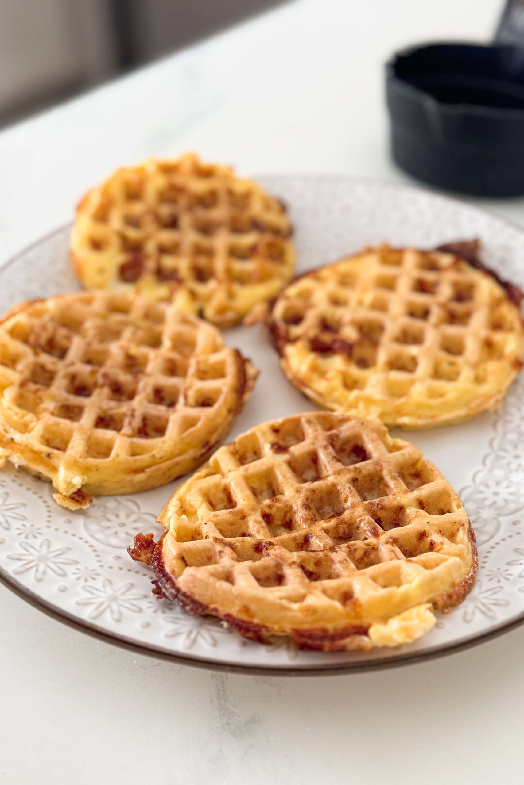 Four golden-brown cheese and egg chaffles rest on a decorative white plate, crispy around the edges. It's a yummy carnivore diet breakfast option.