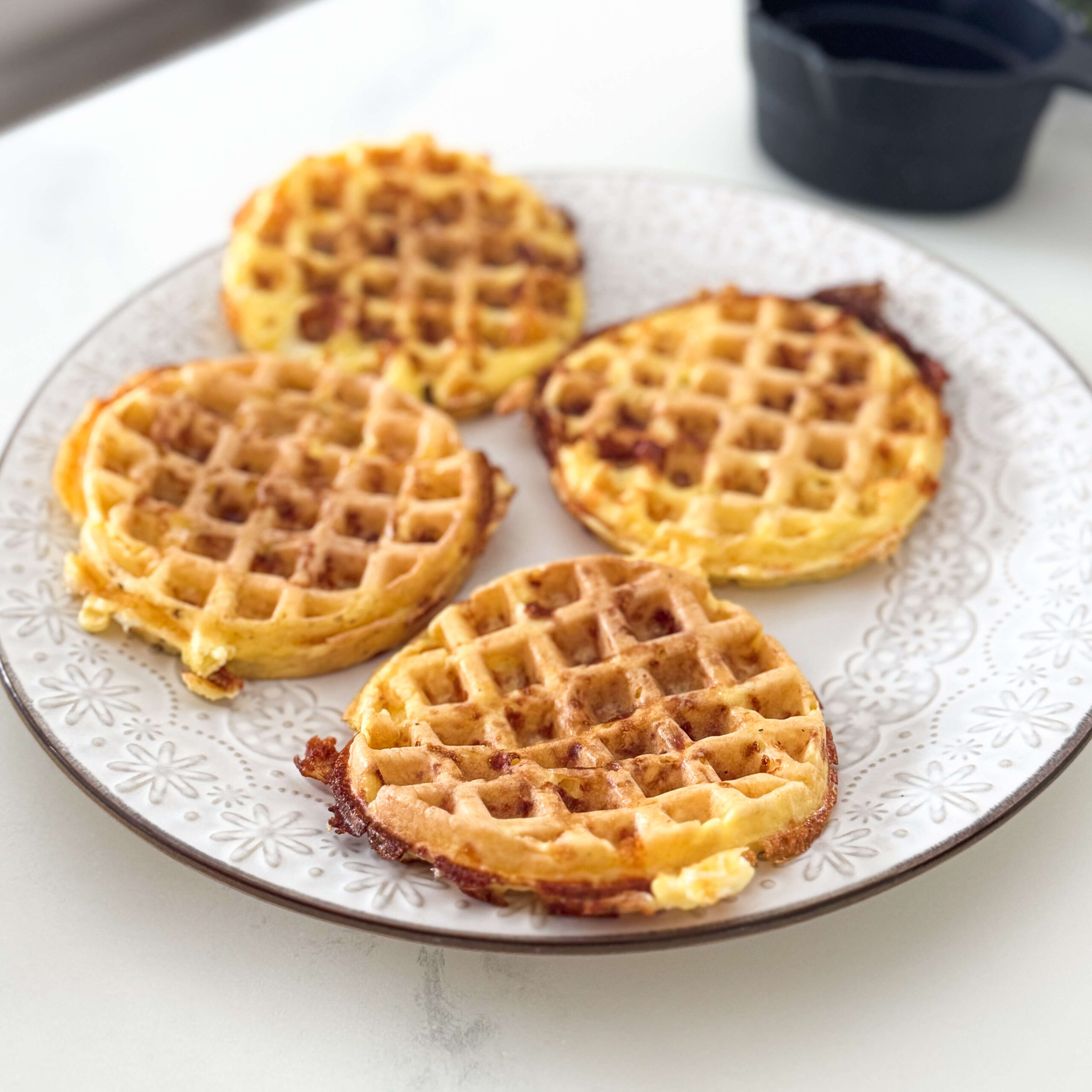 Four golden-brown cheese and egg chaffles rest on a decorative white plate, crispy around the edges. A small black measuring cup sits in the background on a white kitchen counter.