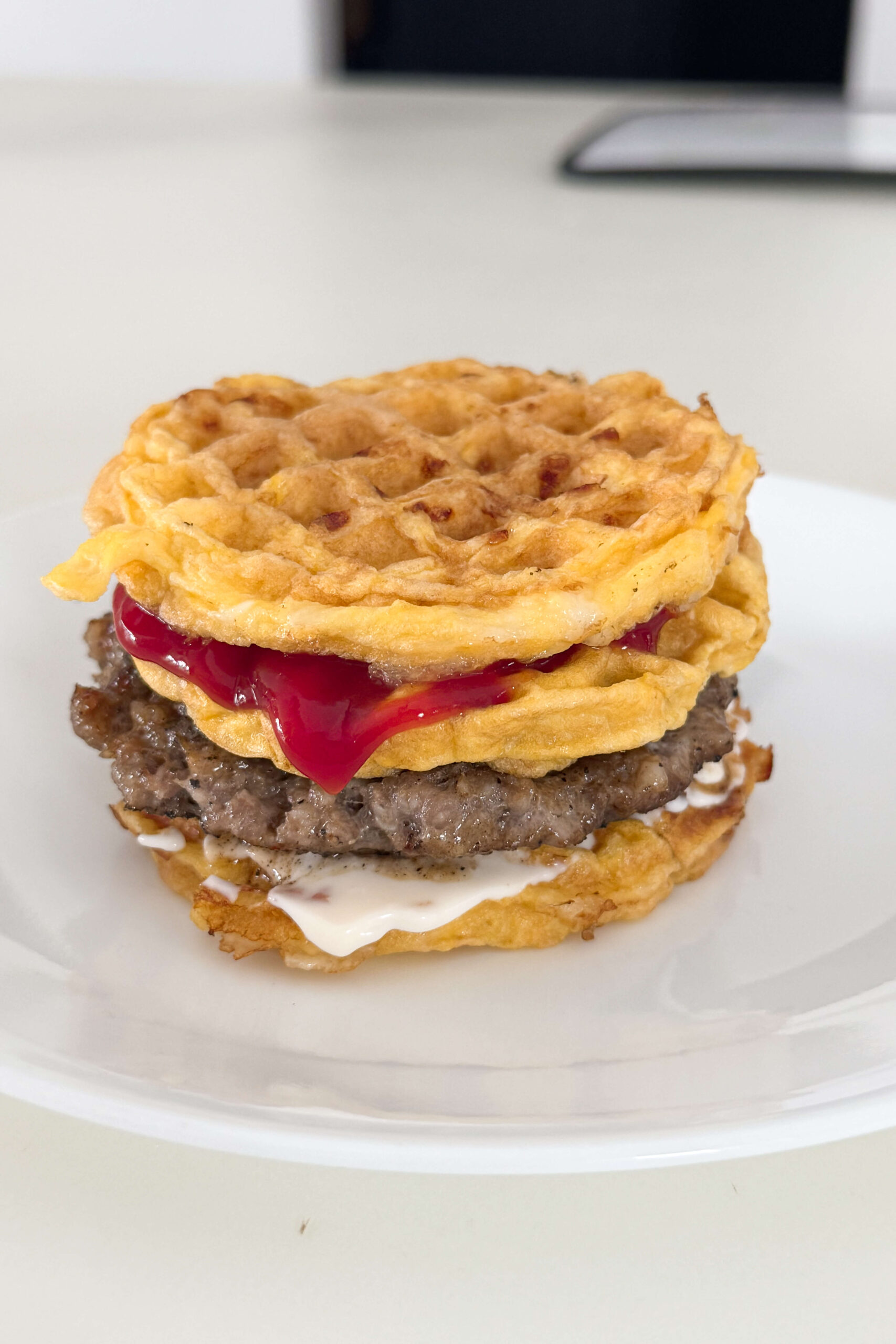 A homemade, high protein breakfast, featuring a chaffle breakfast sandwich with a Jimmy Dean sausage patty, mayonnaise, and ketchup sandwiched between two crispy cheese waffles, served on a white plate.