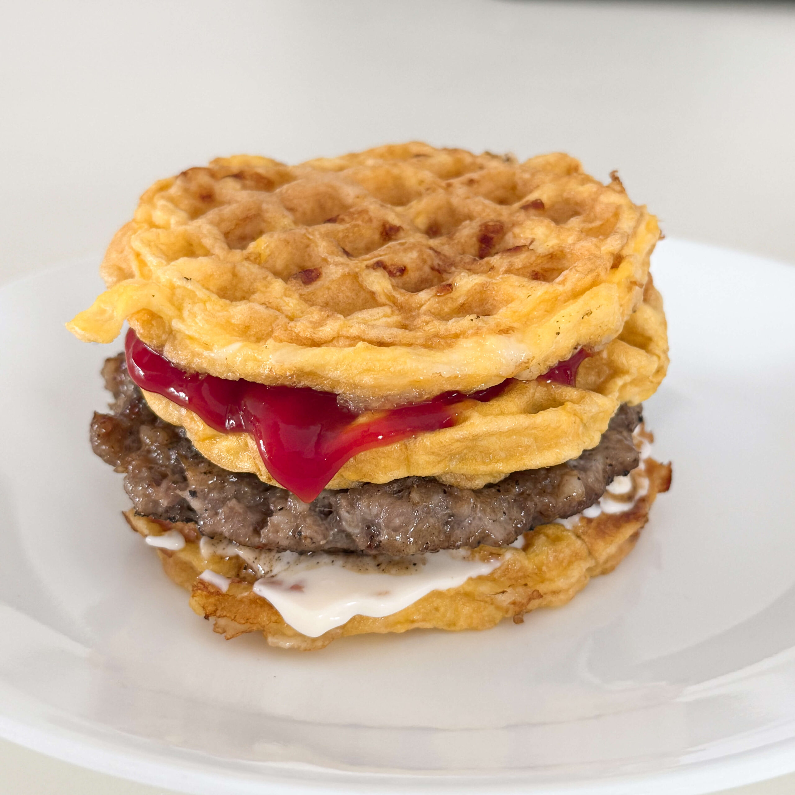 A homemade, high protein breakfast, featuring a chaffle breakfast sandwich with a Jimmy Dean sausage patty, mayonnaise, and ketchup sandwiched between two crispy cheese waffles, served on a white plate.