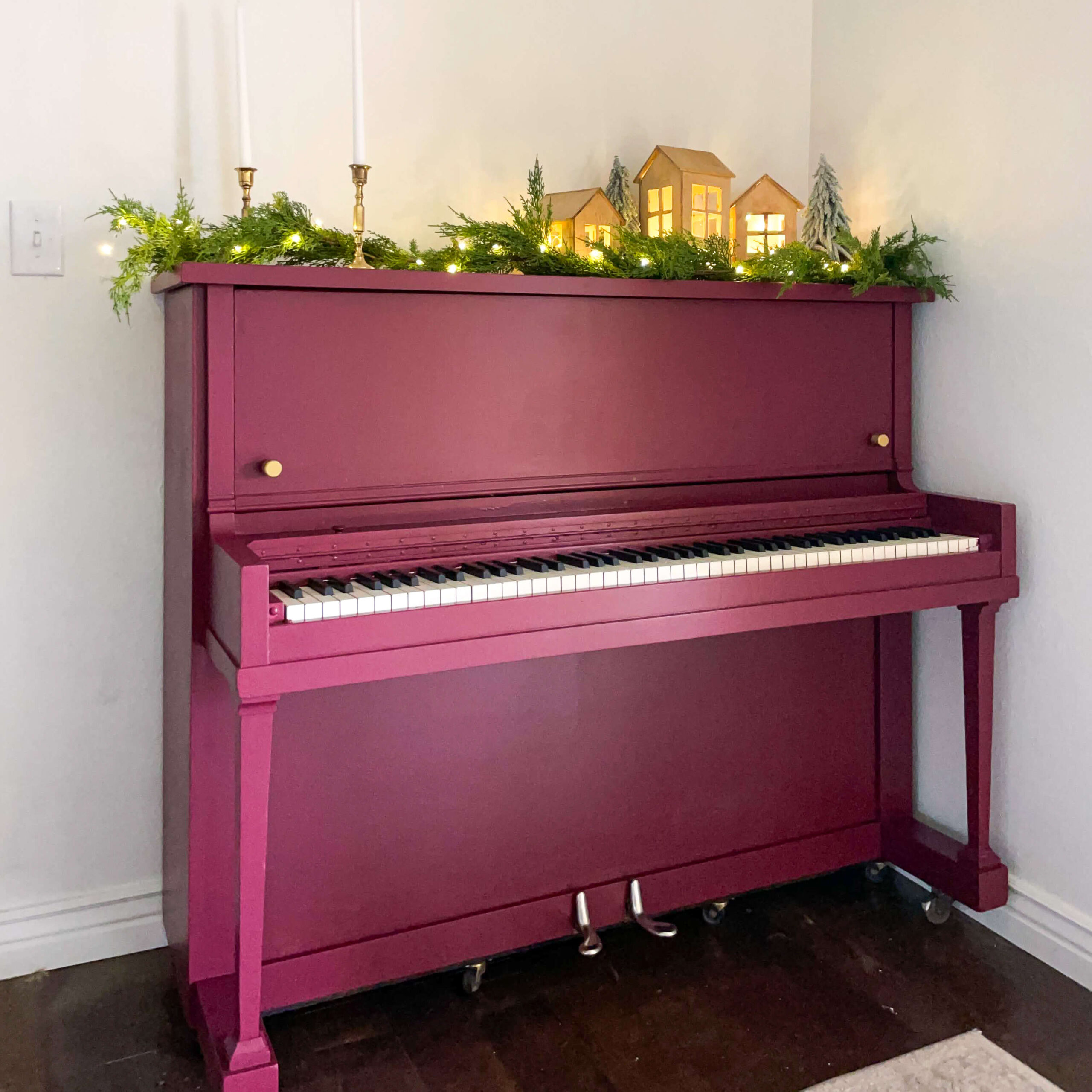 Beautifully painted burgundy upright piano decorated for Christmas with glowing wooden houses, greenery, and brass candlesticks on top.