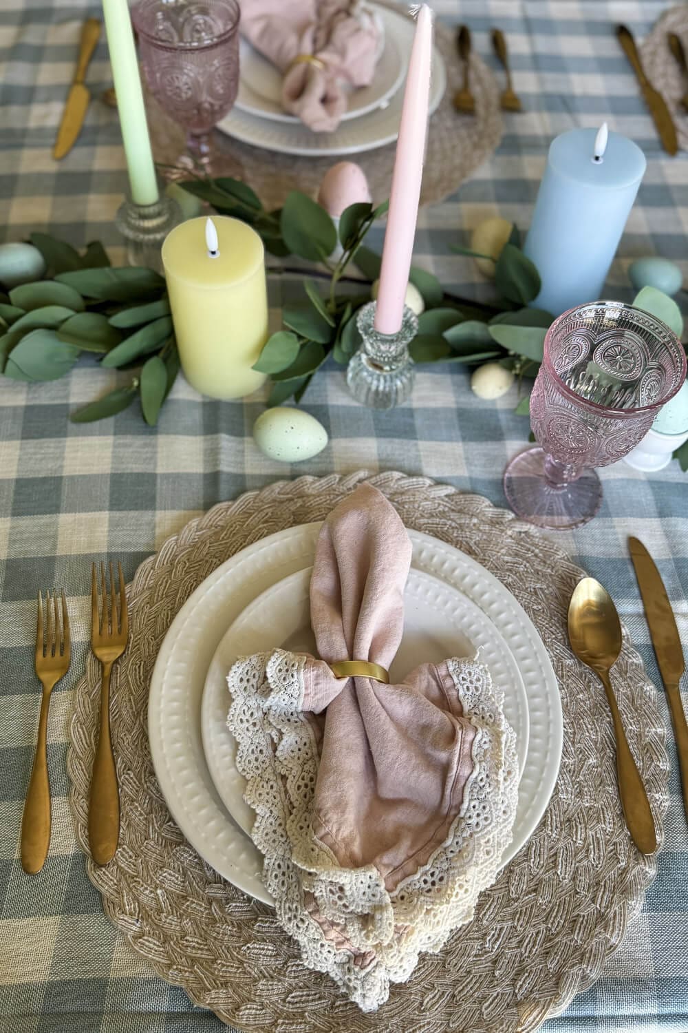 Layered Easter tablescape with woven placemats, white dinnerware, gold flatware, and pink vintage goblets