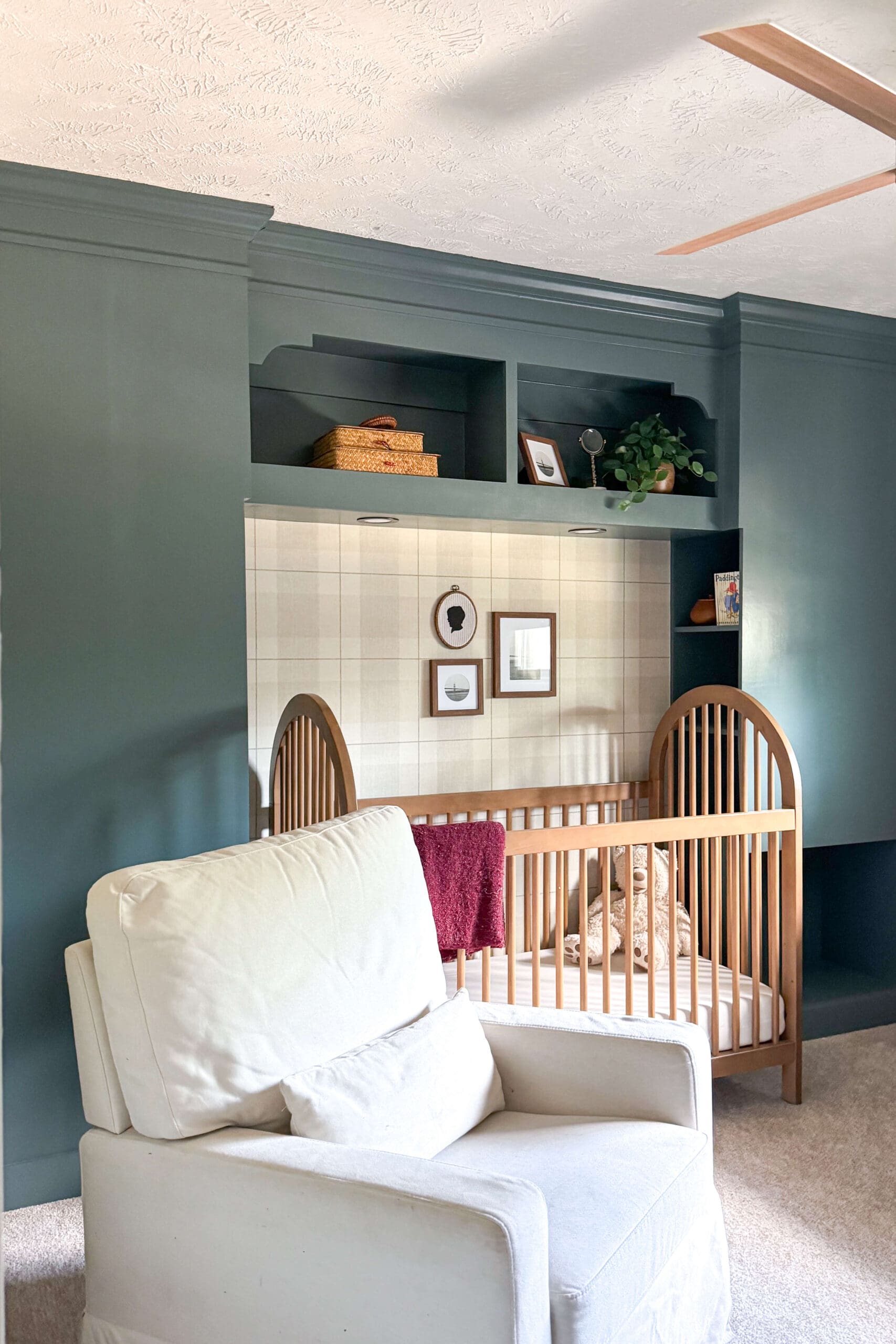 Built-in headboard with storage shelves, painted deep green with a wooden crib, plaid wallpaper accent, and a cozy cream glider in the foreground.