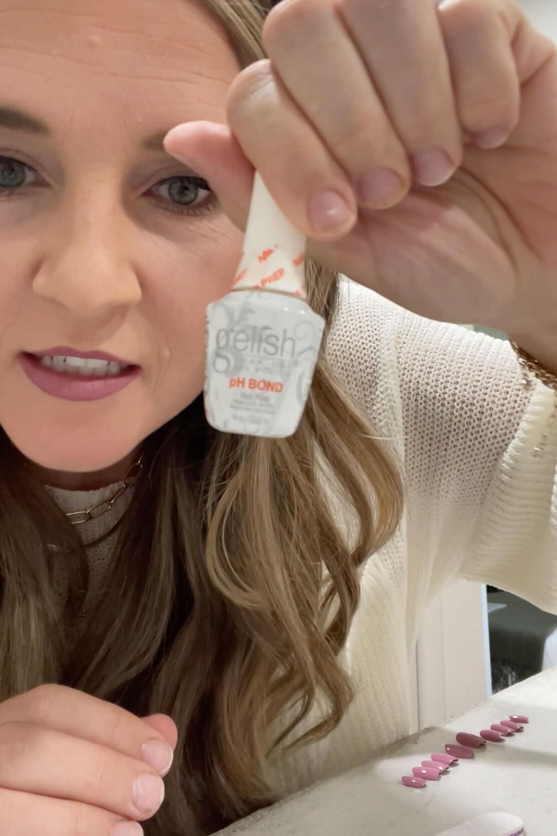 Holding up a bottle of Gelish pH Bond nail prep, an optional extra step to help Kiss Impress press-on nails last longer.