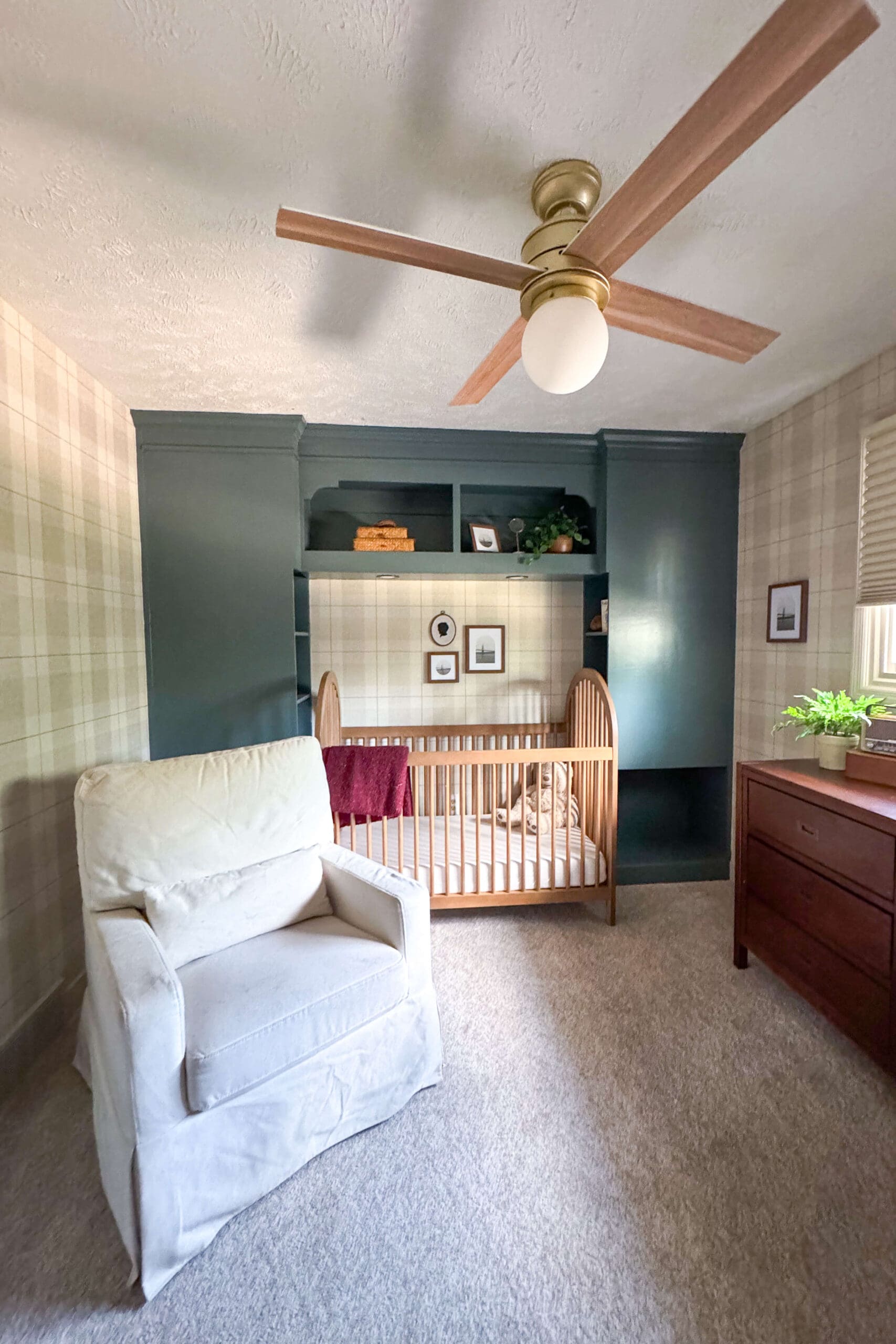 Wide view of nursery with built-in headboard painted deep green, a light wood crib, plaid wallpaper, and modern ceiling fan with wood blades.