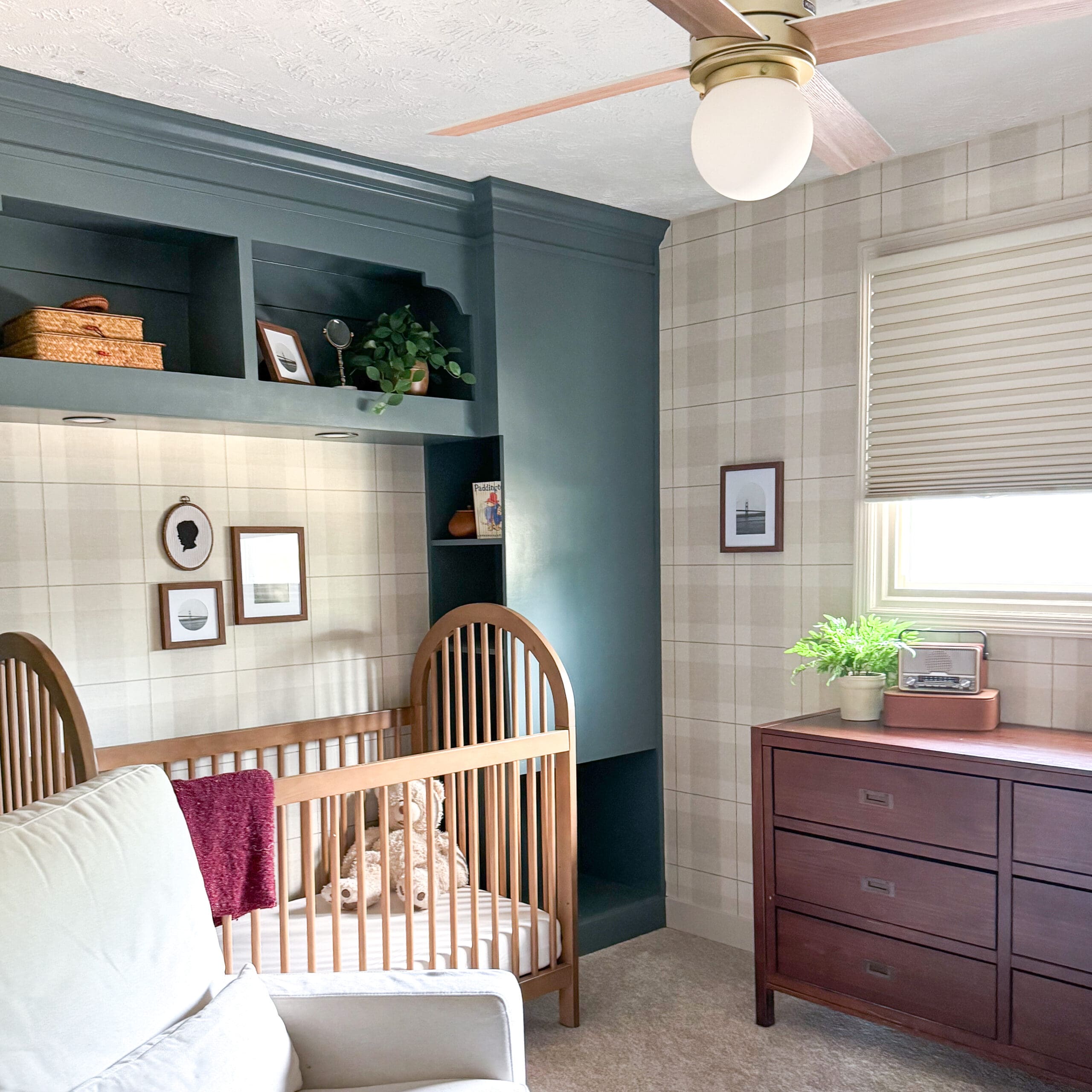 Nursery scene featuring a wooden crib framed by a built in headboard with storage, painted deep green with open shelves, under a plaid wallpaper accent wall and next to a wooden dresser with a potted fern and vintage radio.