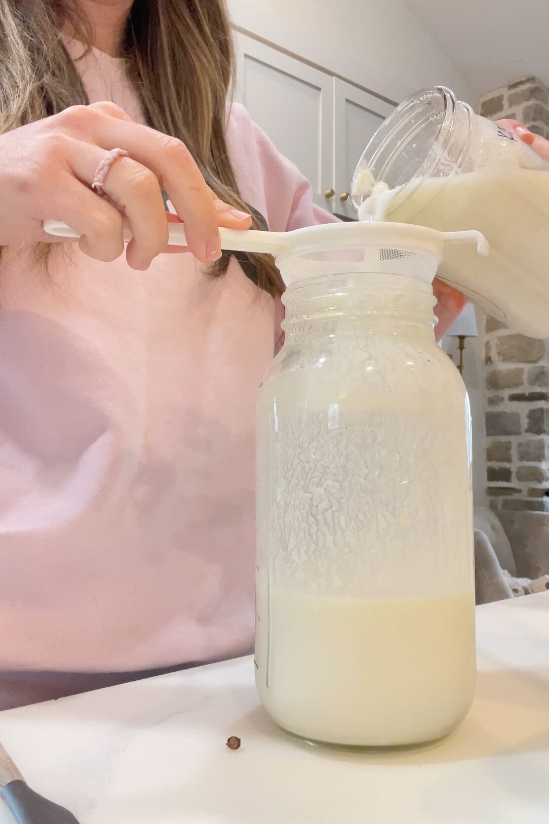 Pouring freshly fermented milk kefir through a plastic mesh strainer into a clean jar while showing how to make milk kefir at home.