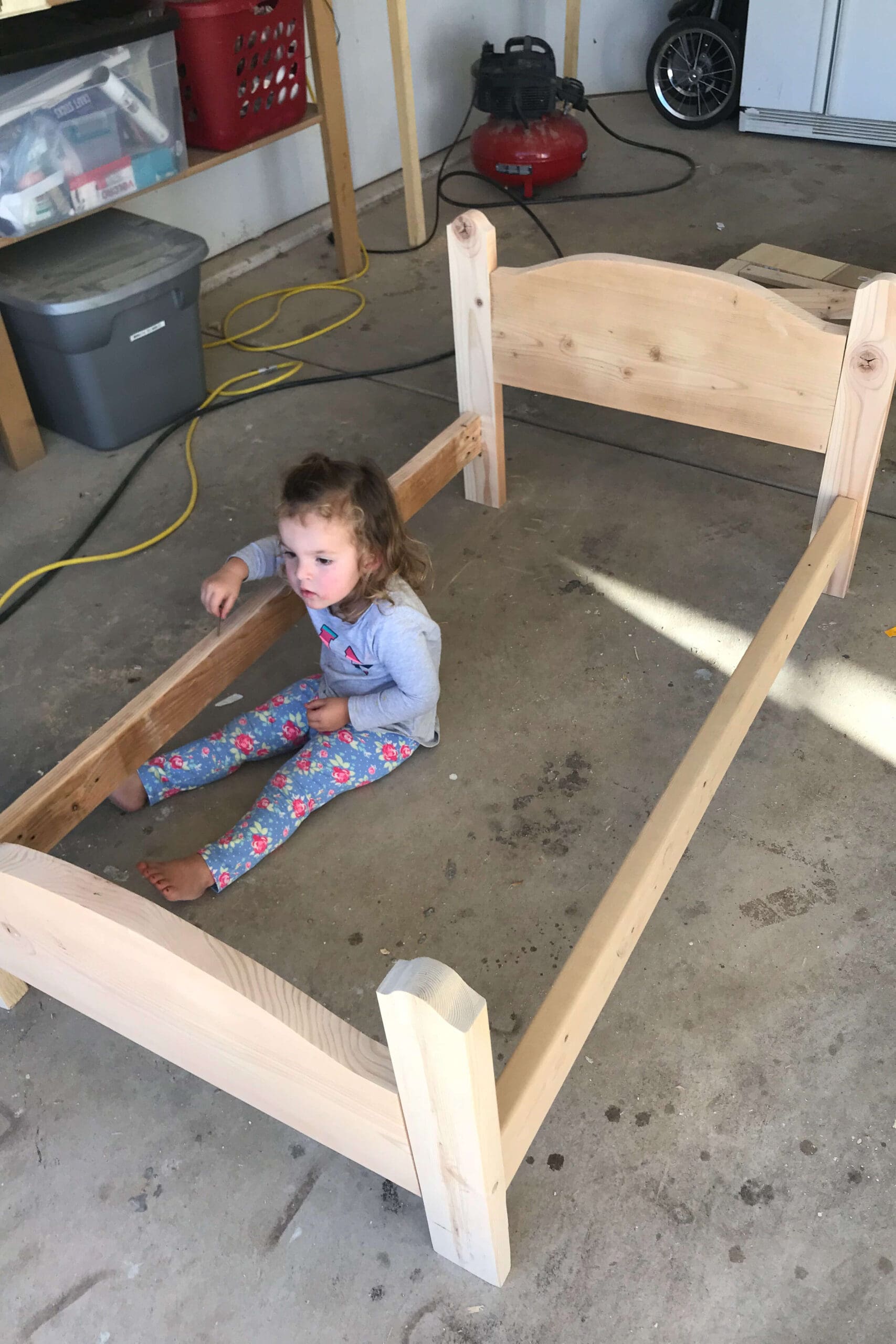 Partially assembled wooden bed frame in a garage with a young child sitting inside, helping during the DIY build process.