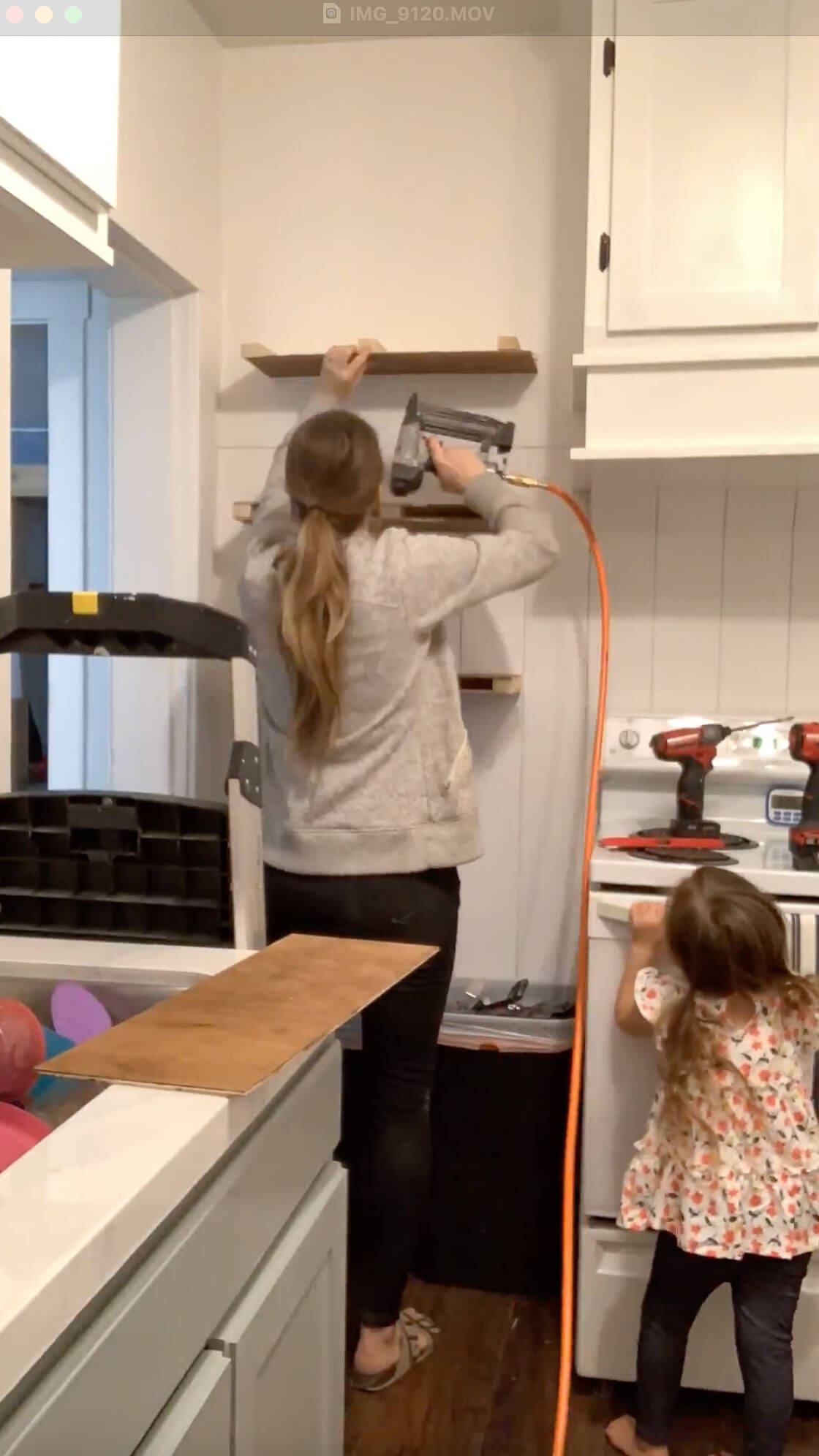 Installing the plywood top of a DIY floating shelf using a brad nailer, with a toddler nearby and tools staged on the stove—part of a family-friendly kitchen DIY project.
