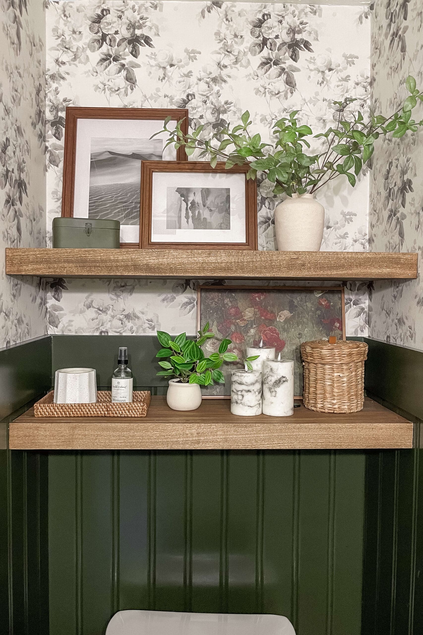 Two DIY floating shelves styled in a floral wallpapered bathroom, featuring wood tones, framed art, greenery, and cozy accessories for a custom built-in look.