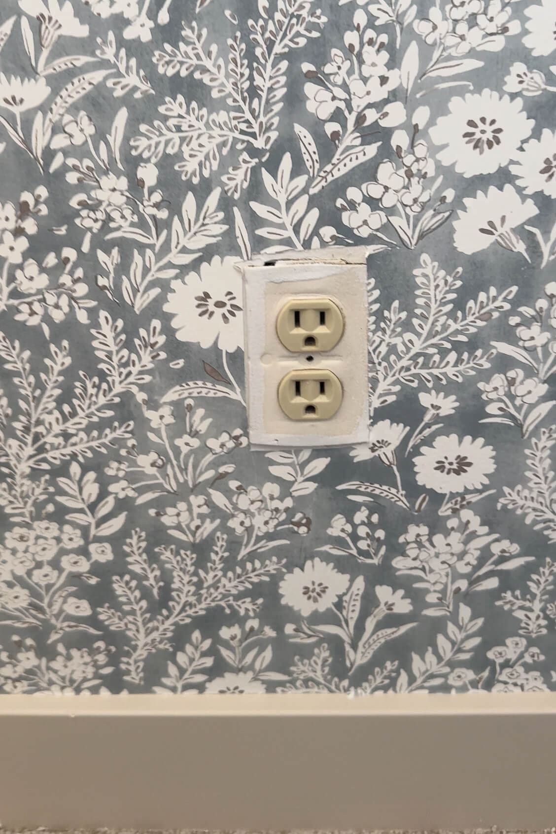 Standard white outlet on a blue floral wallpapered wall, showing the unfinished look before adding a camouflaged cover.