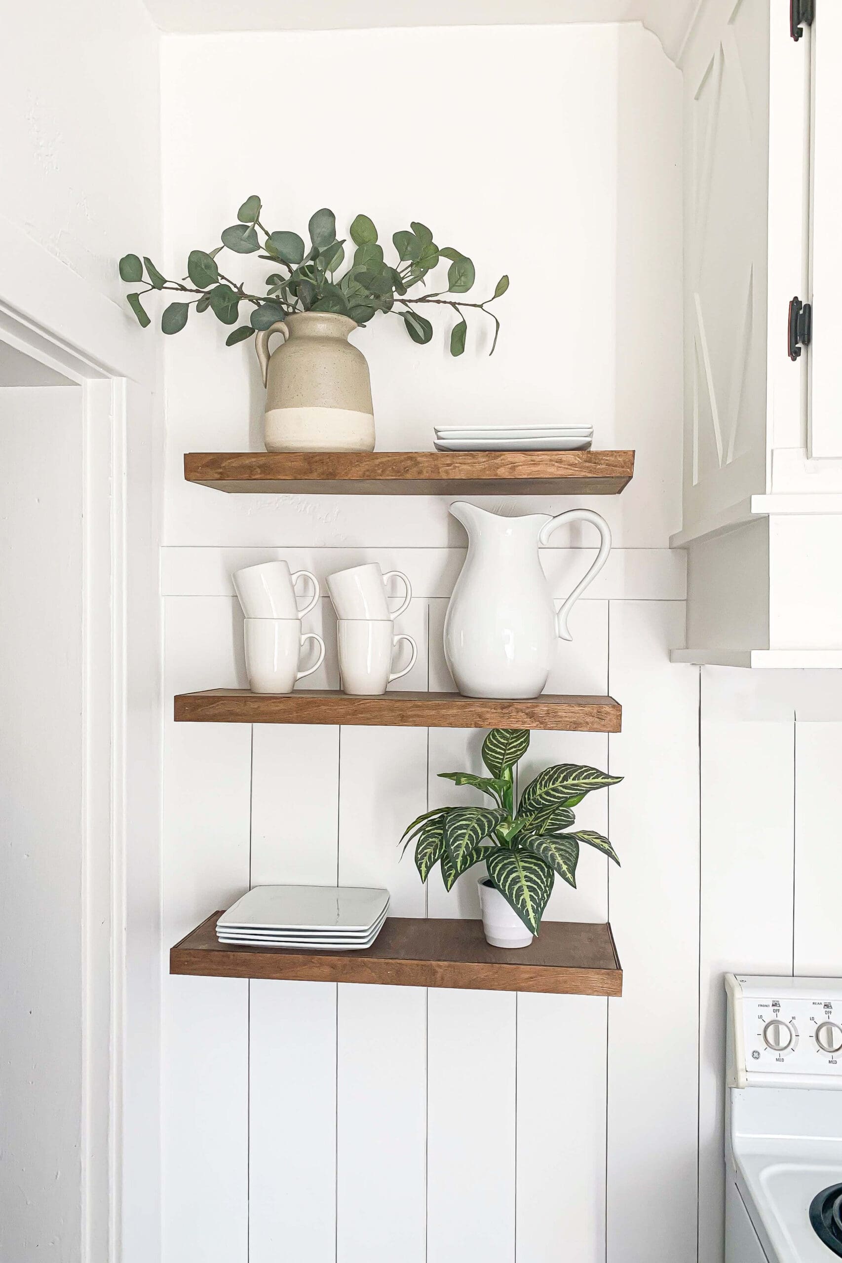 Straight-on view of handmade DIY floating shelves installed without brackets, featuring wood tones against a white kitchen wall, styled with dishes and a ceramic jug for a clean, modern farmhouse look.