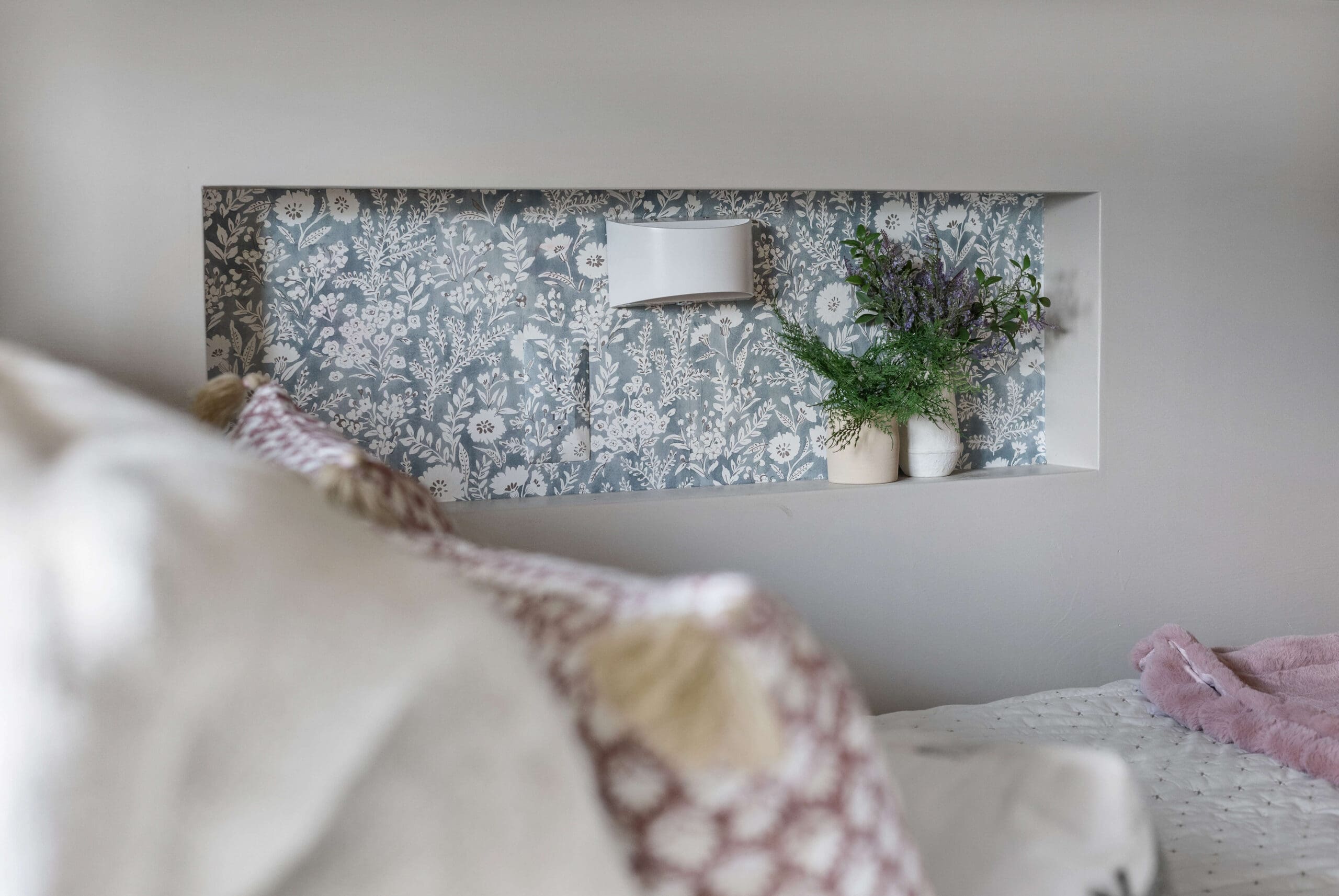 Styled wall niche with soft blue floral wallpaper and a nearly hidden outlet cover, decorated with potted greenery for a cozy built-in look.