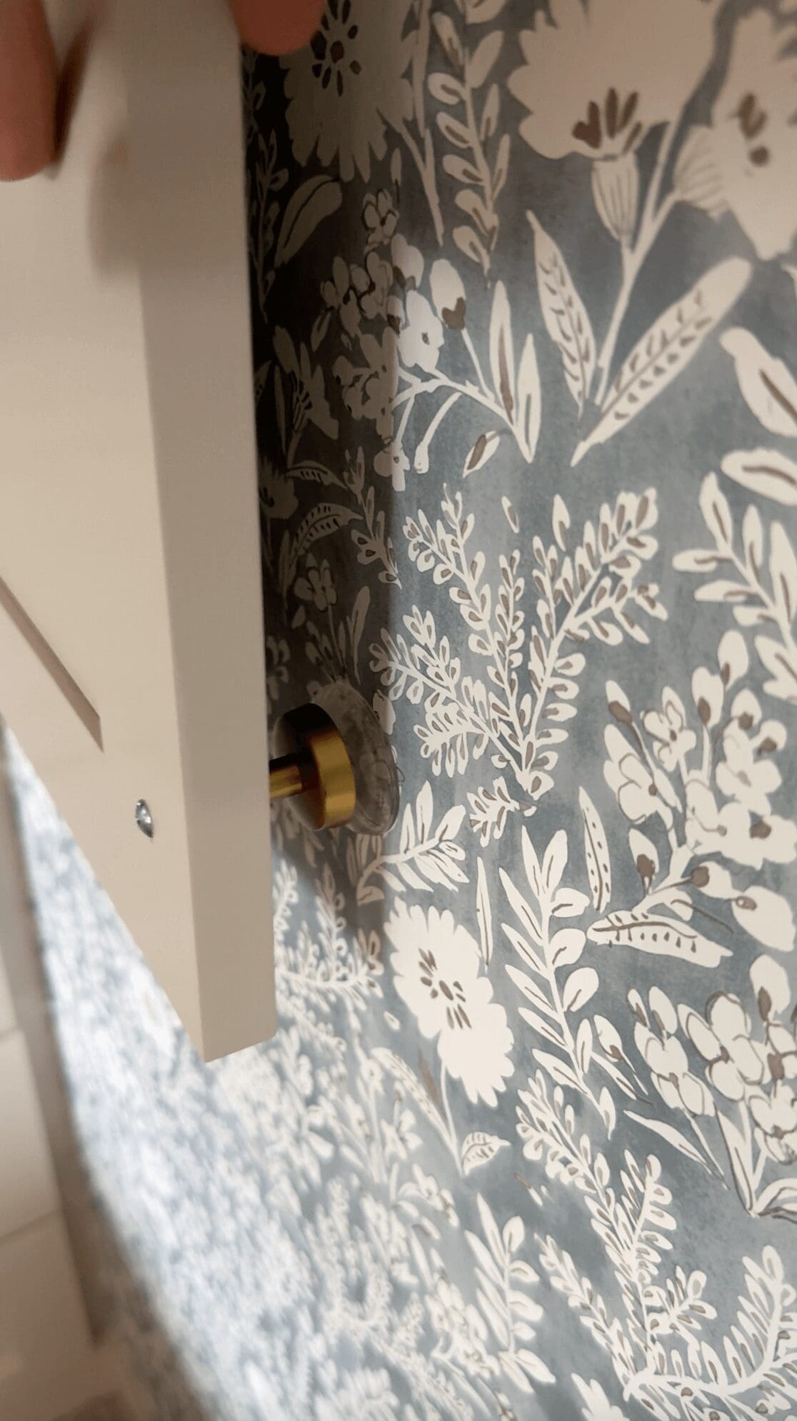 Gold cabinet knob resting against a floral wallpapered wall with a clear adhesive bumper applied to prevent damage.
