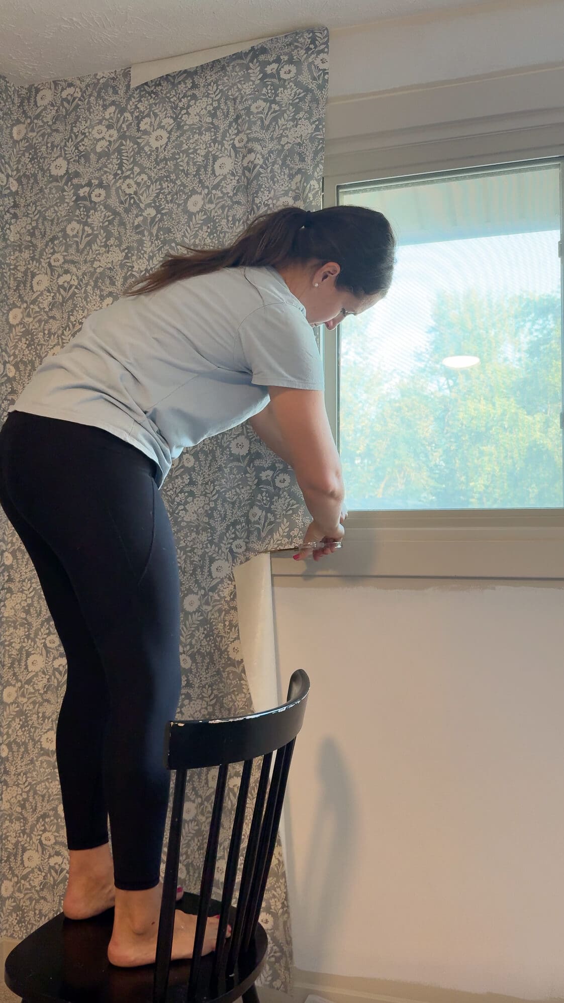 Carefully trimming wallpaper along the bottom edge of a window frame while standing on a chair during installation.