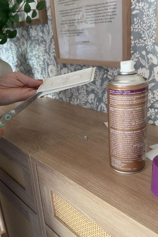 Trimming excess wallpaper from the back of a switch plate using scissors, with a can of adhesive nearby on the workspace.