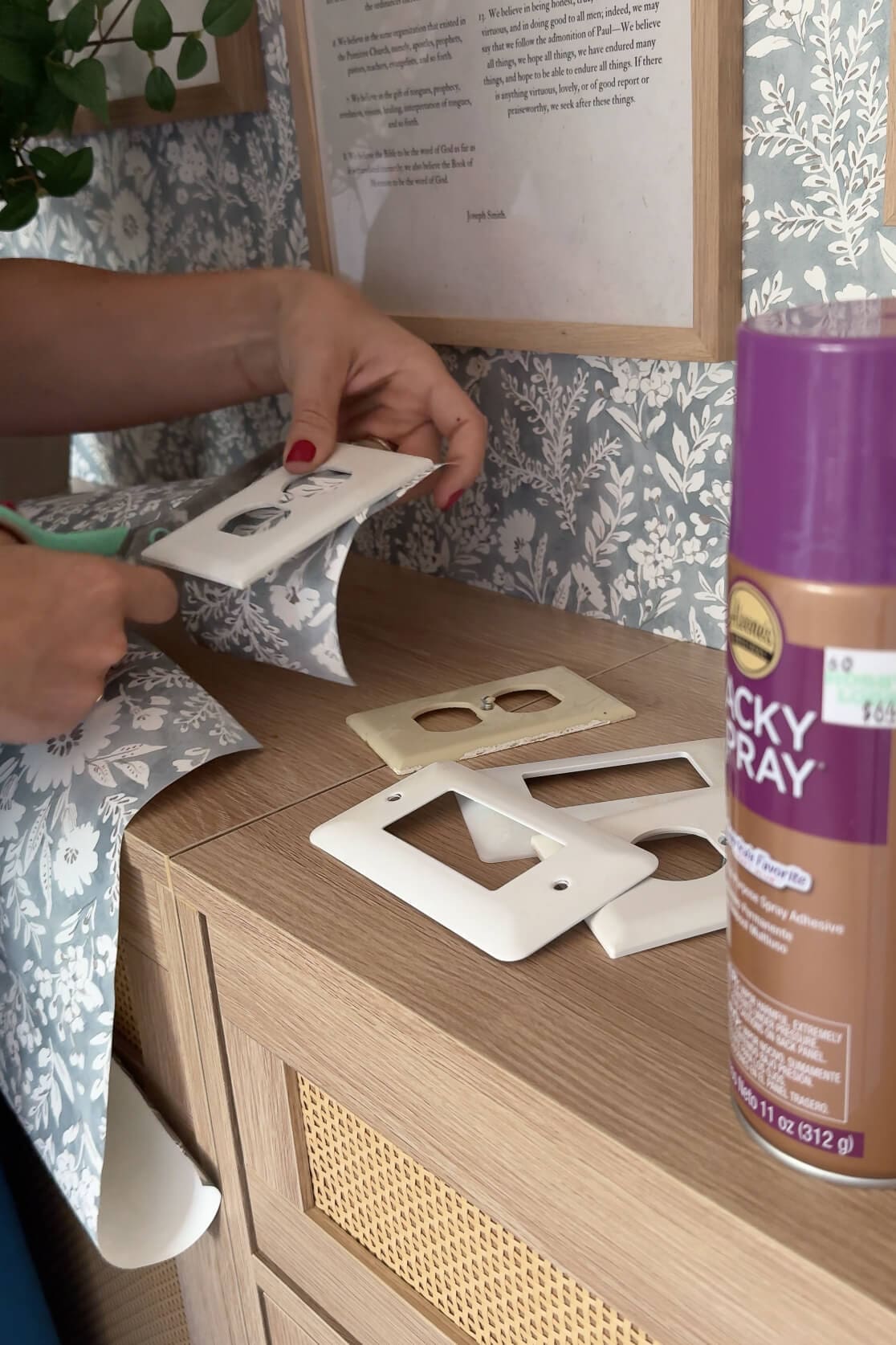 Hands cutting floral wallpaper to size around a white wall plate, mid-DIY project.