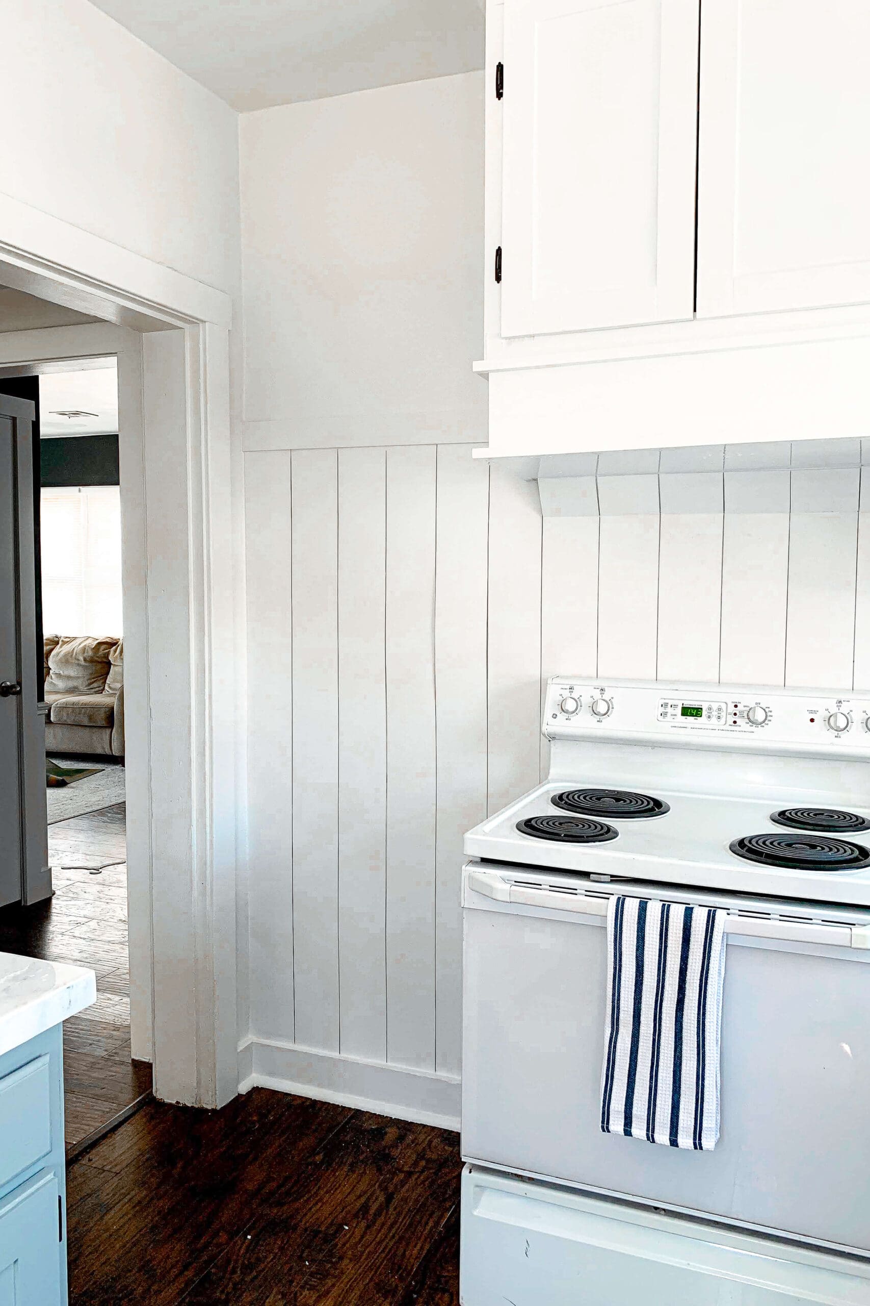White shiplap accent wall in a bright kitchen corner with upper cabinets and a white stove, part of a budget-friendly DIY kitchen renovation project.