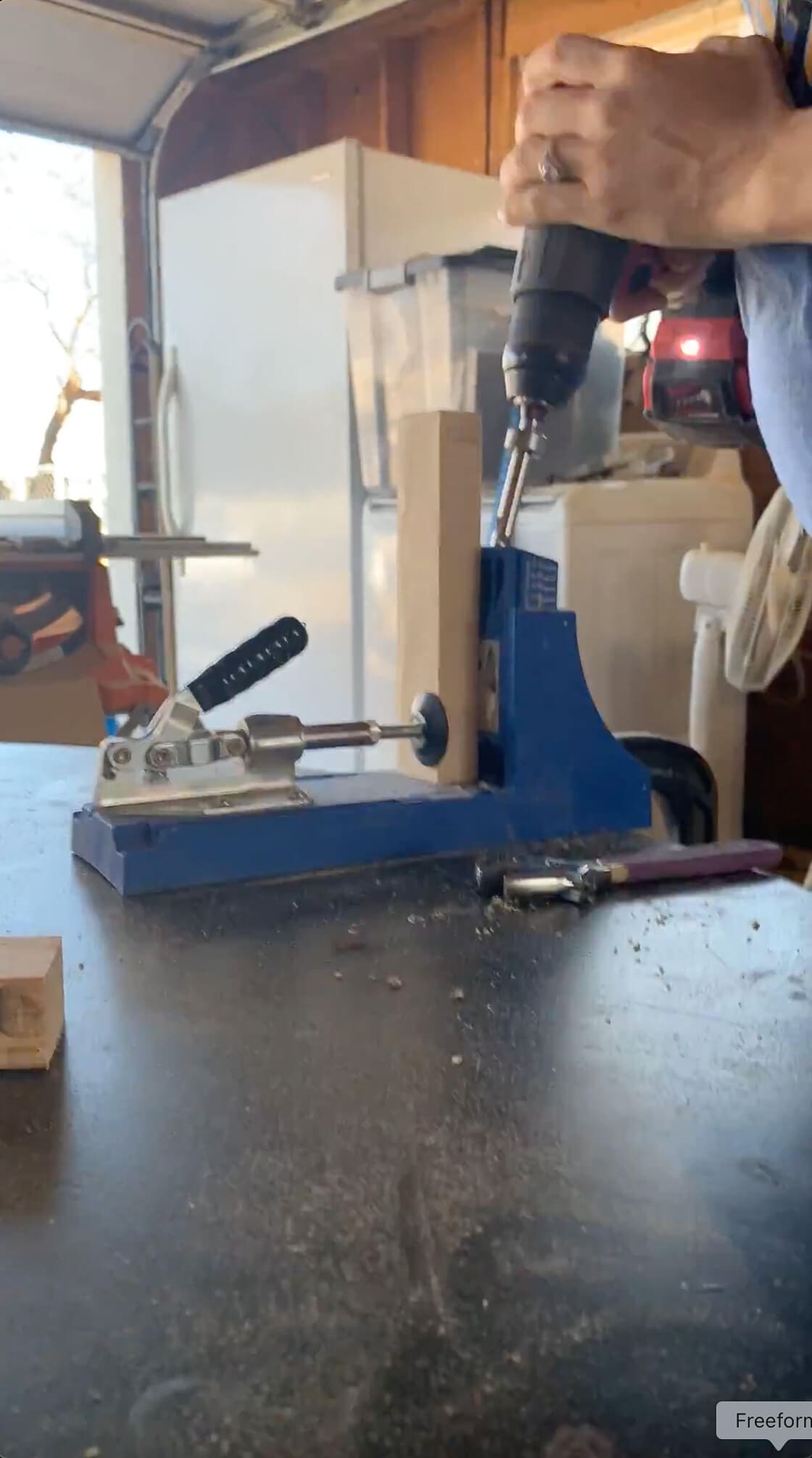 Using a drill to create pocket holes in a wooden board with a Kreg jig, part of a DIY woodworking project in a home garage workspace.

