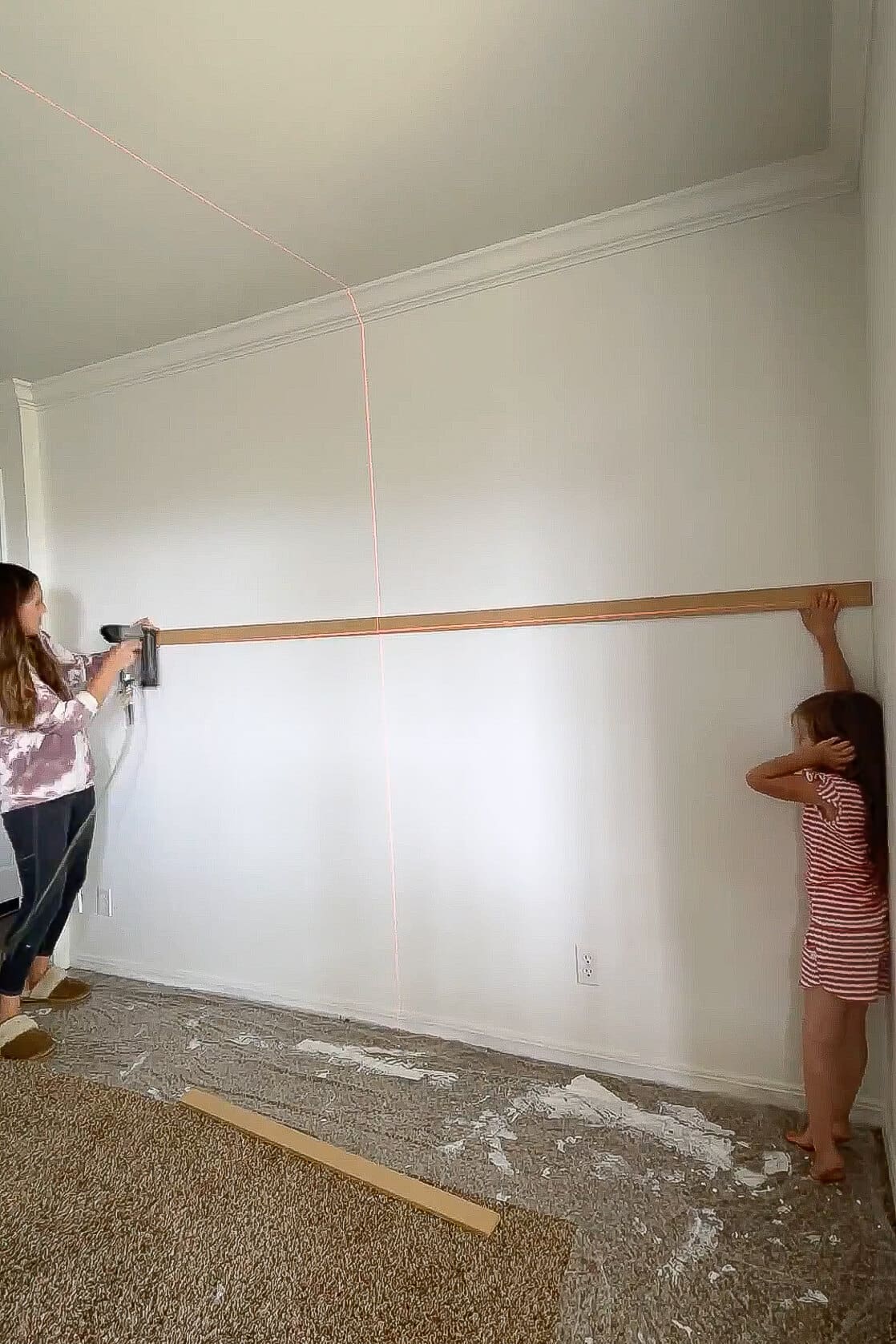 Mom installing a horizontal trim piece with a nail gun while her daughter helps hold it steady during a family-friendly DIY project.