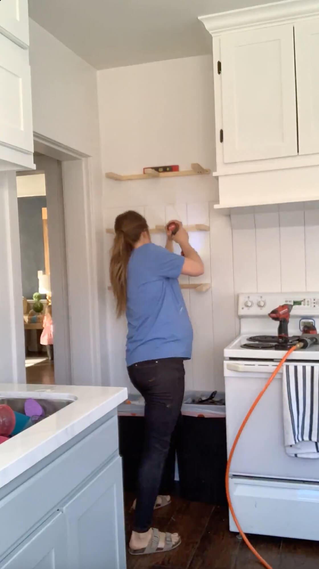 DIYer using a power drill to install shelf frames onto a white shiplap wall, part of a floating shelf project in a bright farmhouse-style kitchen.