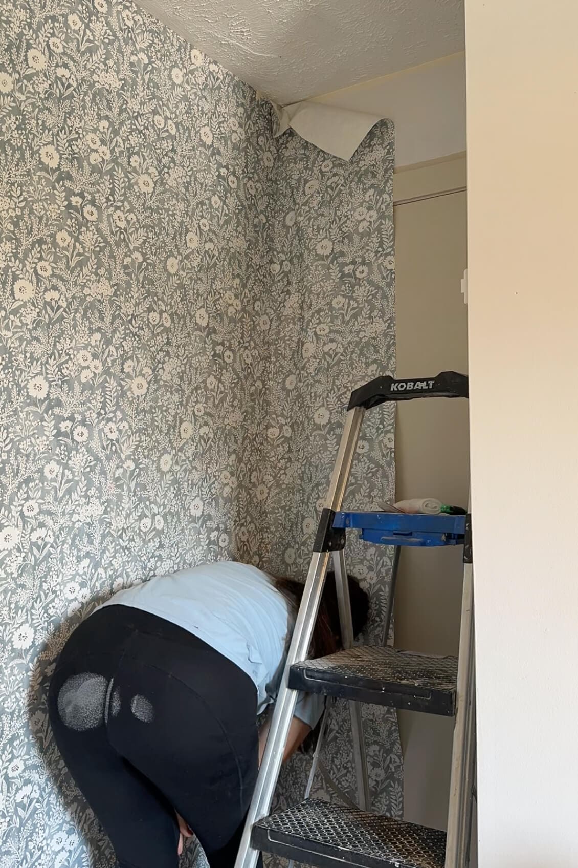 Installing the final wallpaper panel in a tight corner using a ladder, with the floral pattern continuing around the doorway.