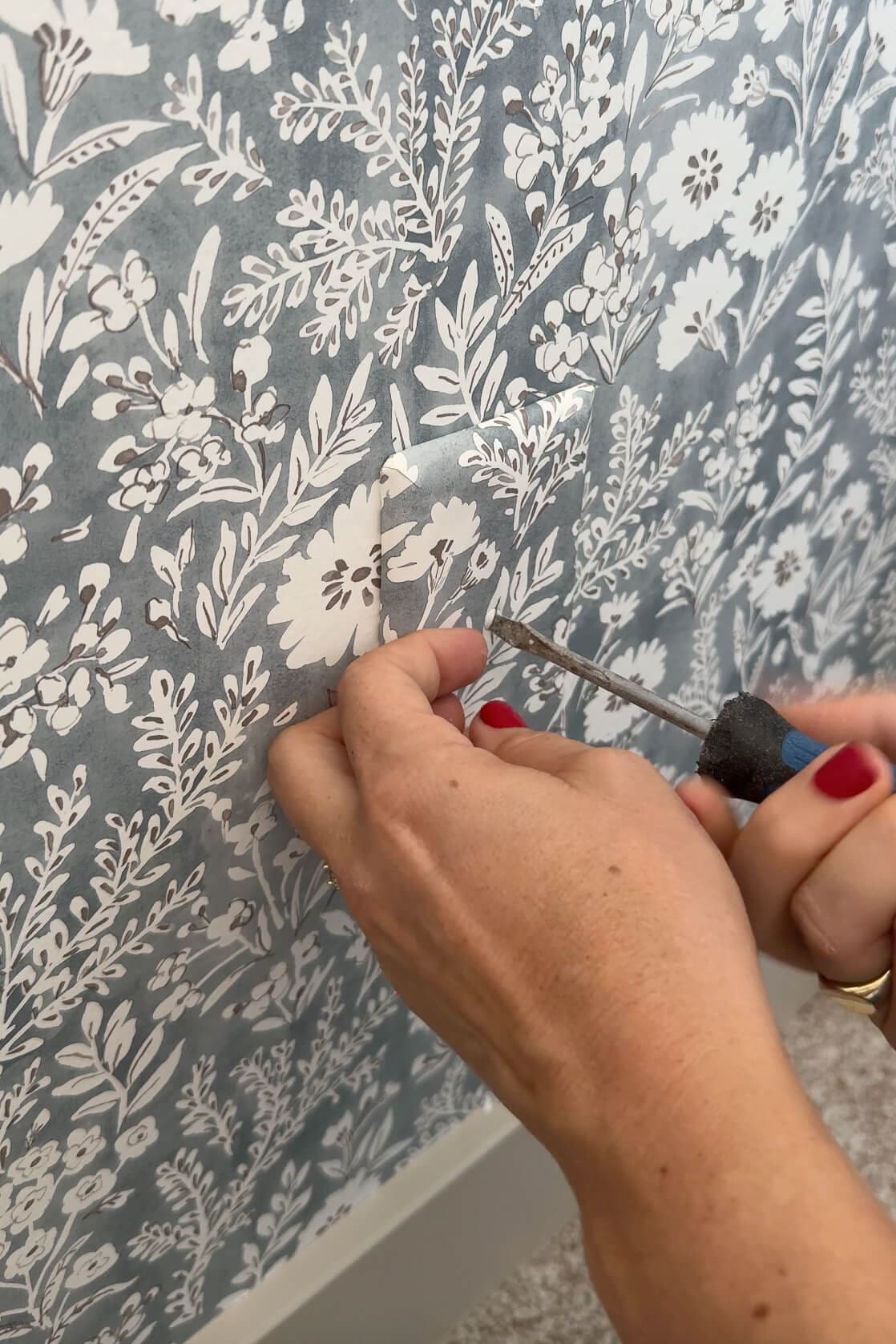 Close-up of hands screwing a wallpapered outlet cover into the wall, perfectly aligned with the floral pattern for a seamless look.
