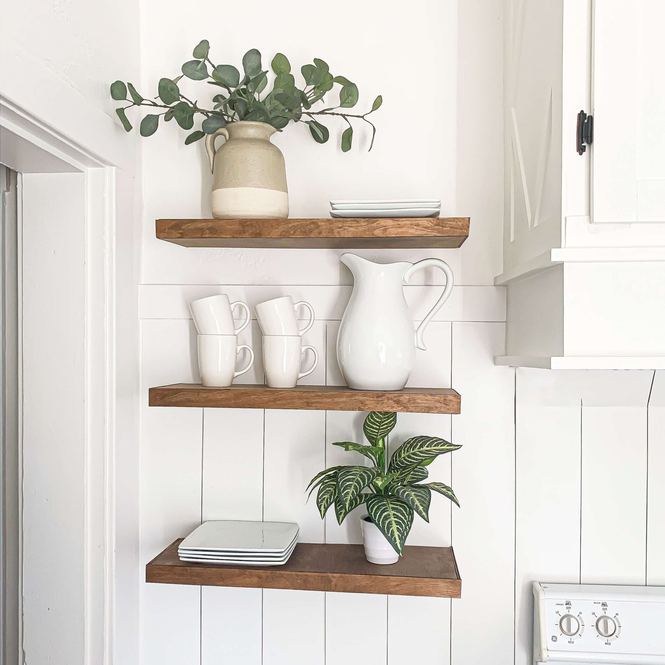 Close-up of three DIY floating shelves in a white shiplap kitchen wall, decorated with white ceramic dishes, a large pitcher, stacked plates, and a leafy potted plant—an affordable and stylish kitchen storage idea.