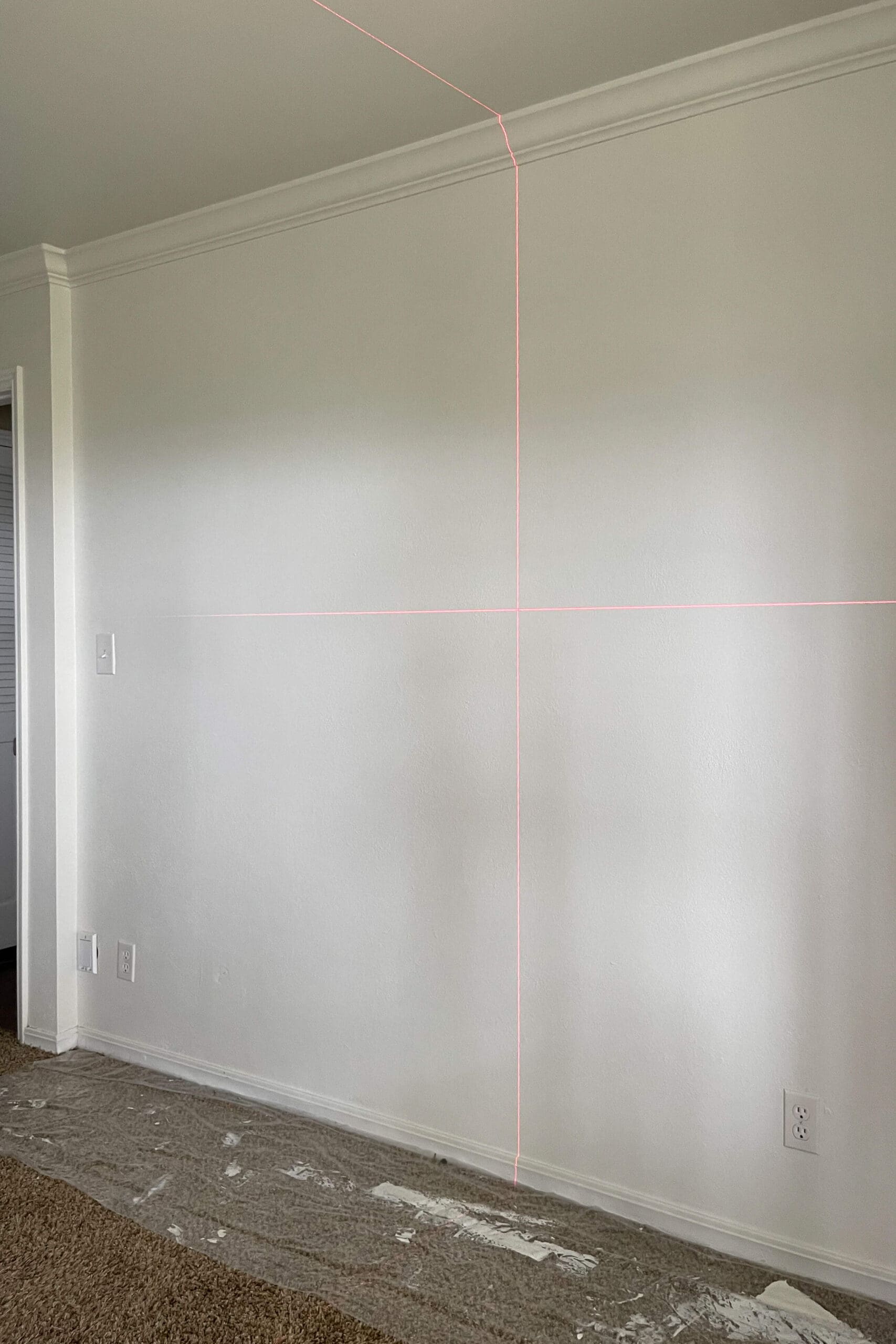 Laser level projecting vertical and horizontal guide lines onto a freshly painted wall to plan a beginner board and batten layout.