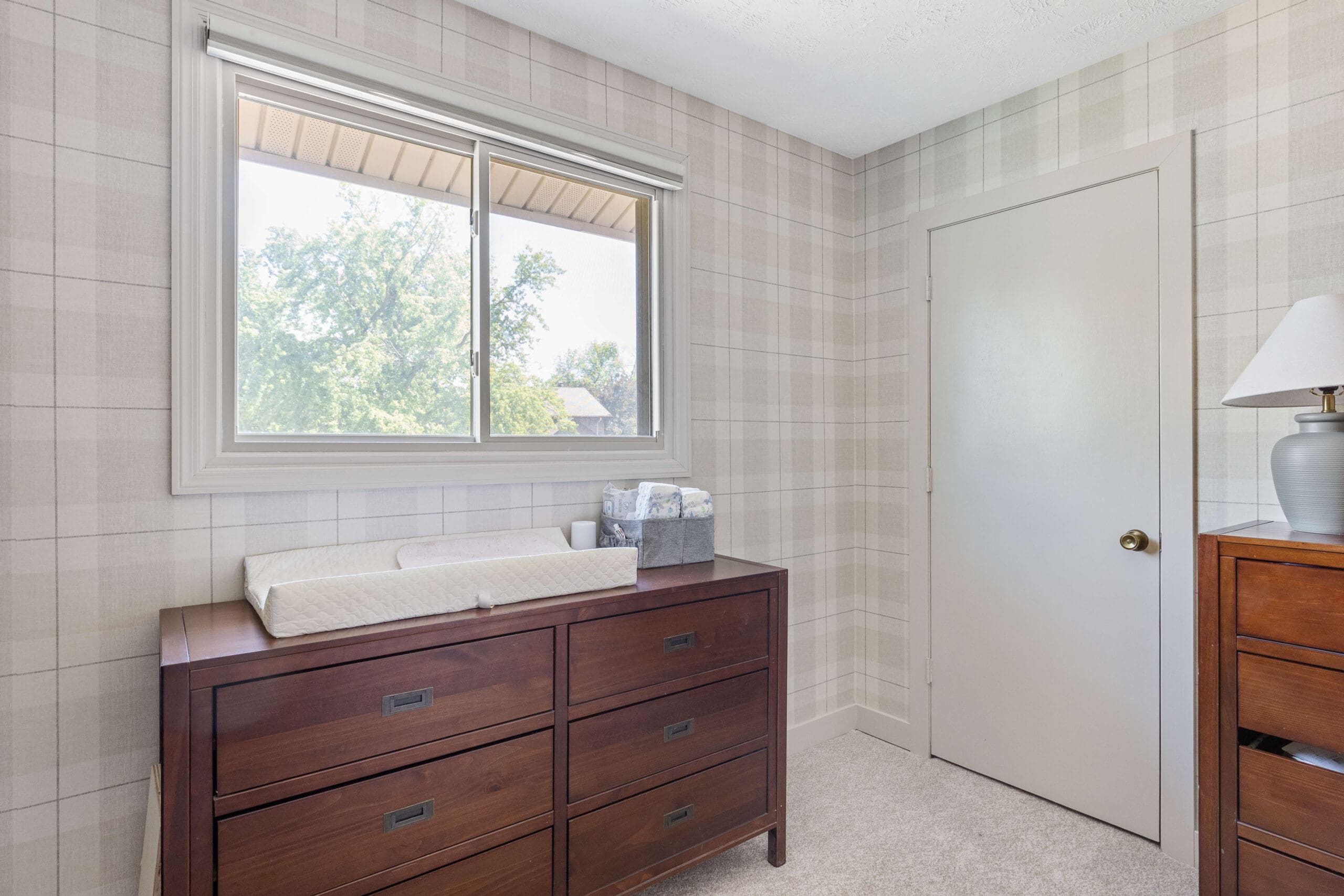 Nursery corner with soft plaid wallpaper, light trim painted in Sherwin Williams Accessible Beige, and rich wood furniture for a cozy, neutral look.