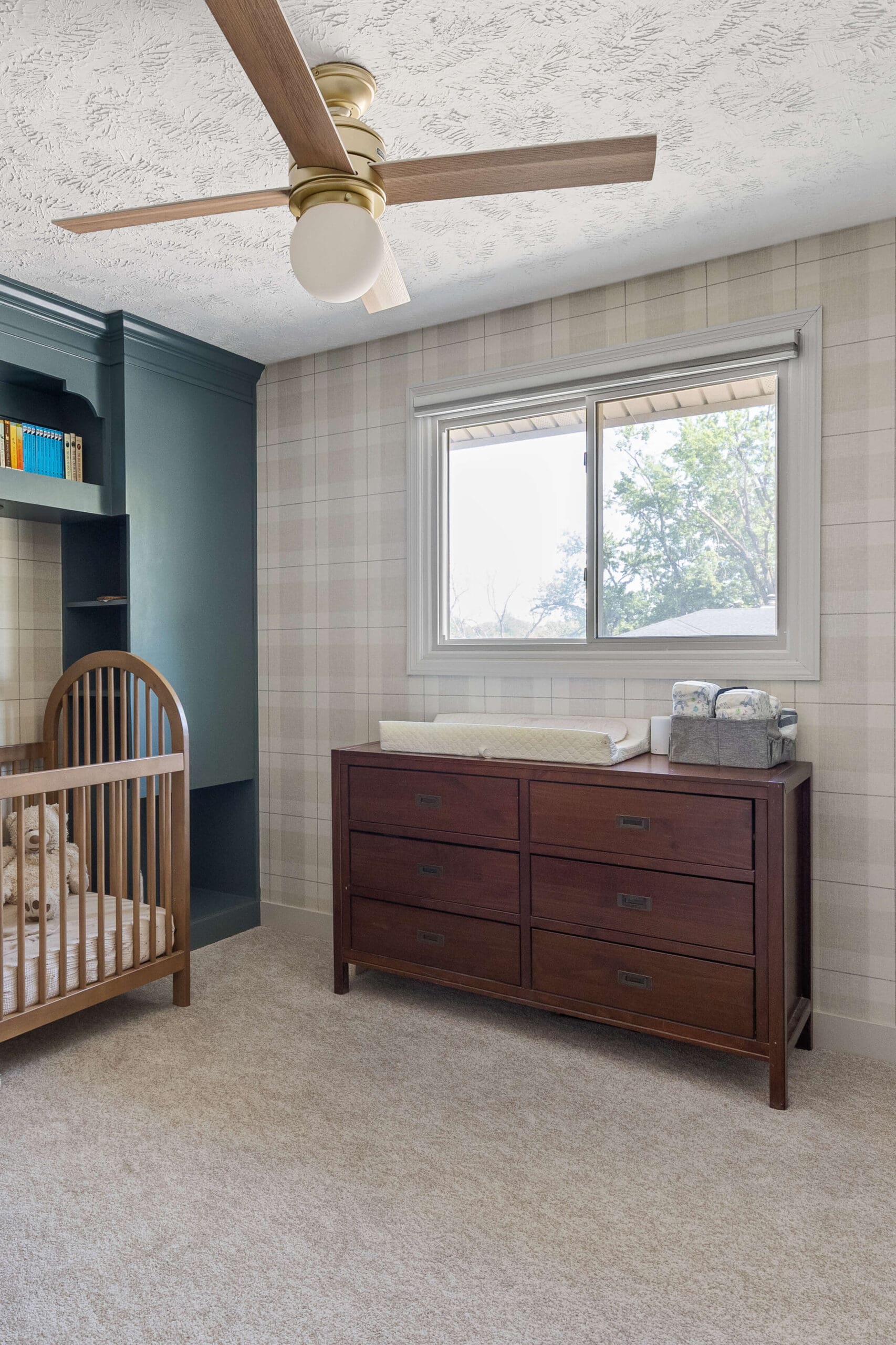 Cozy nursery featuring neutral plaid wallpaper, a wooden crib with stuffed animals, a dark wood dresser with a changing pad and diaper caddy, and built-in sage green shelving next to the crib.