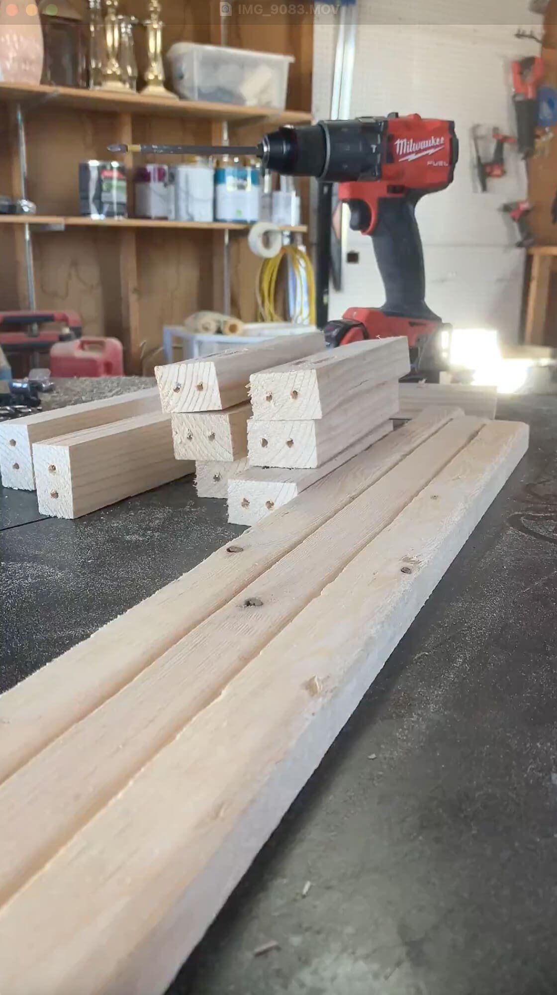 Freshly cut wood boards with pocket holes drilled on the ends, staged on a workbench next to a red power drill in a garage workshop setup.